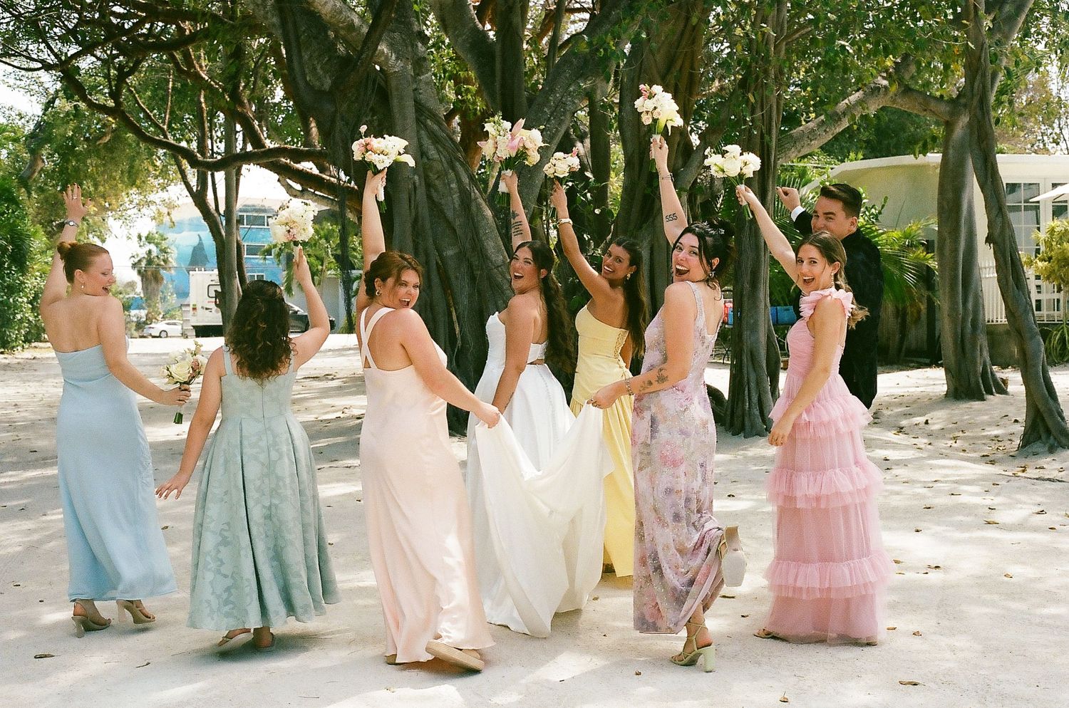Bride and bridesmaids in pastel gowns celebrate outdoors, raising bouquets under tropical trees on sandy ground.