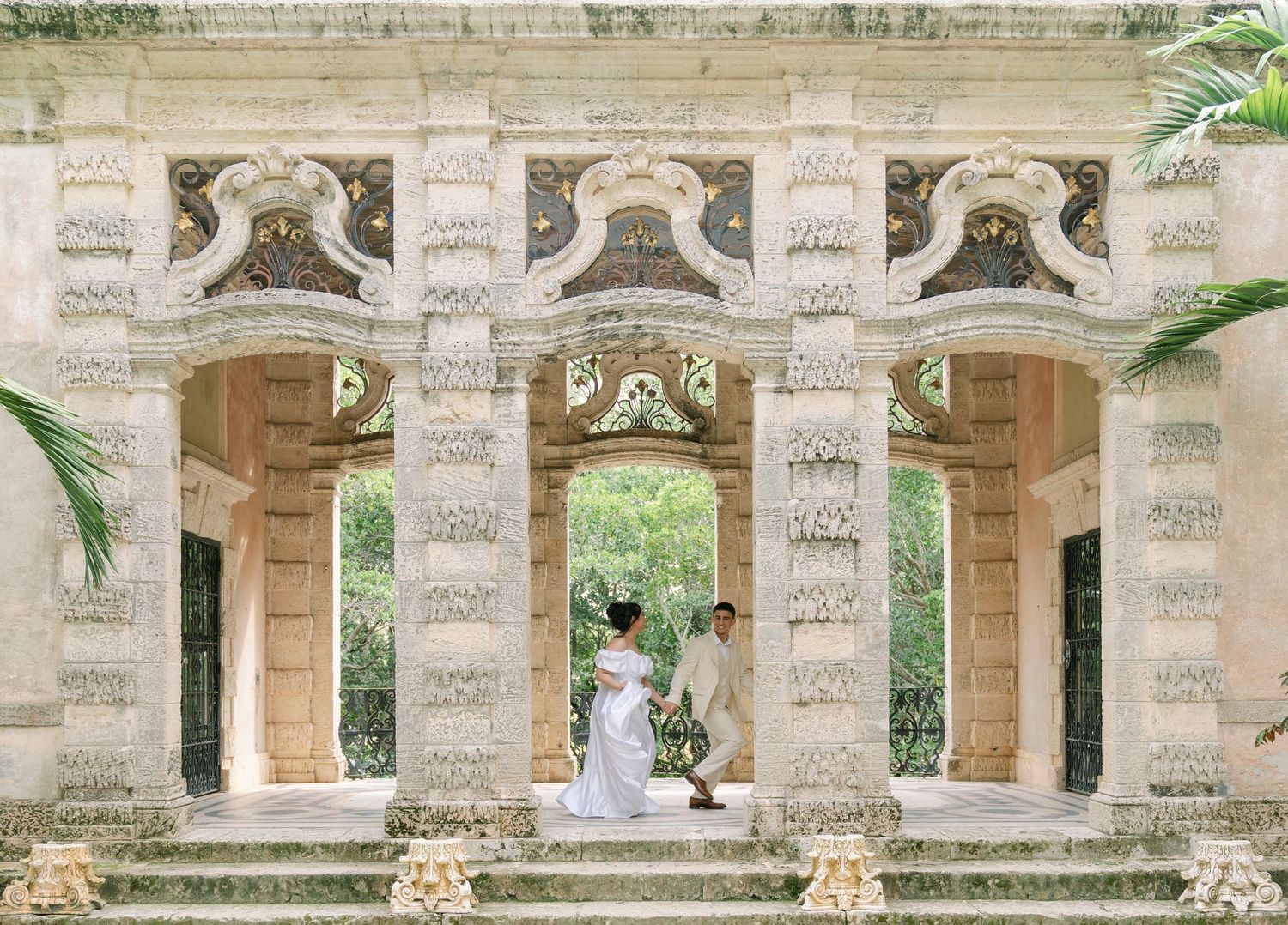 Elegant limestone columns frame a romantic scene at Vizcaya Museum and Gardens in Miami with palm trees visible outside.