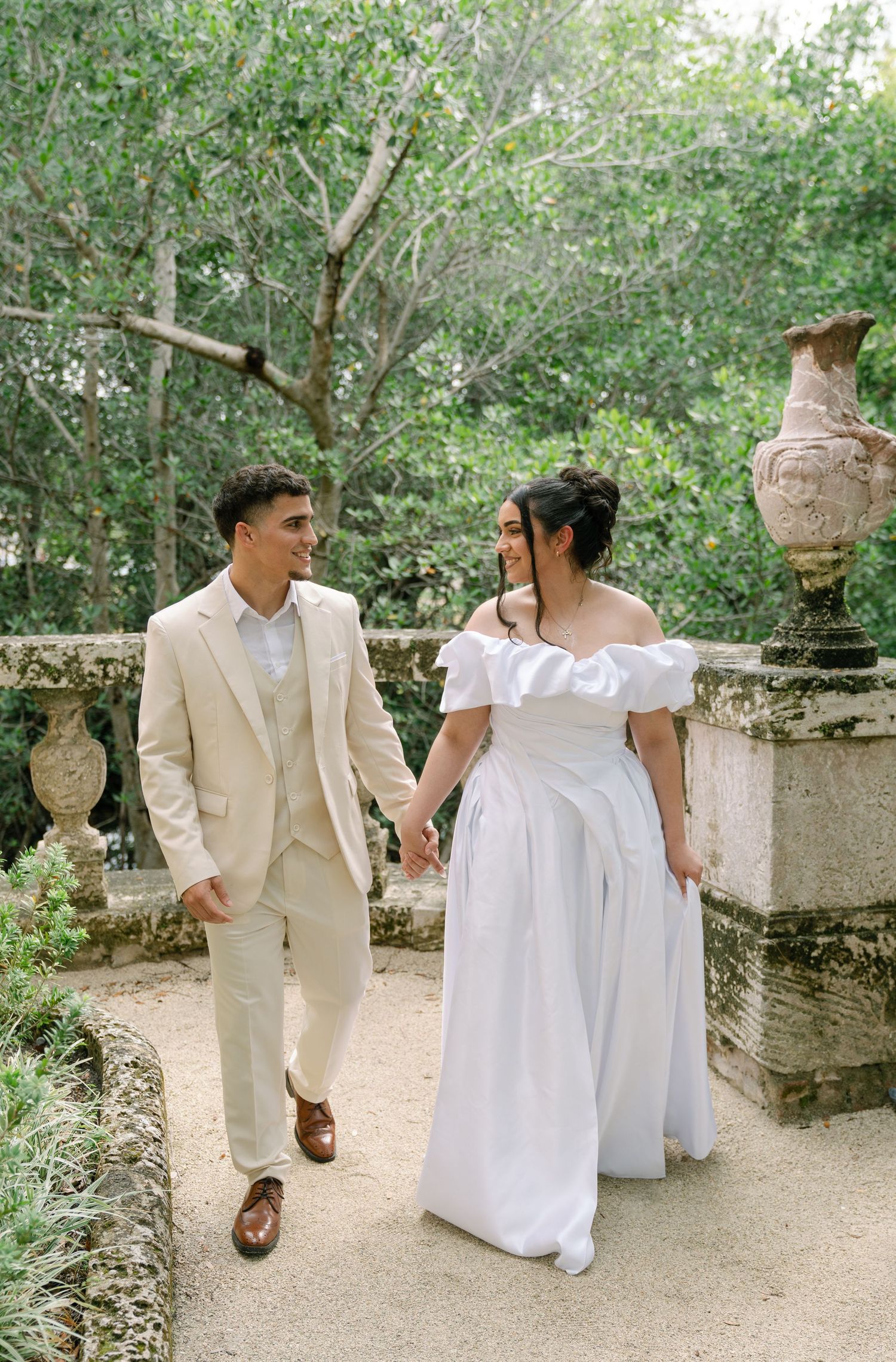 A couple in matching white and cream attire walks hand in hand along a garden path with decorative stone urns.