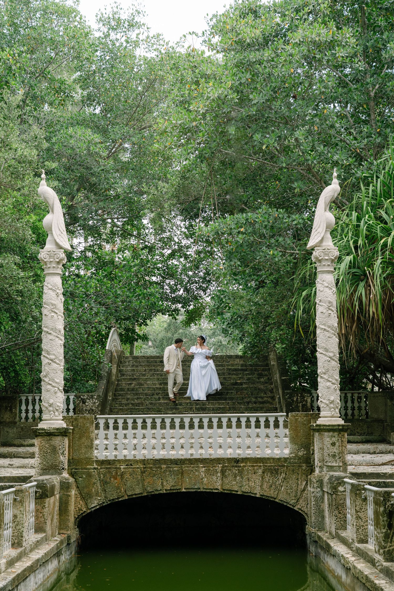 Two people walk along an ornate stone balustrade surrounded by lush greenery in a historic garden setting.