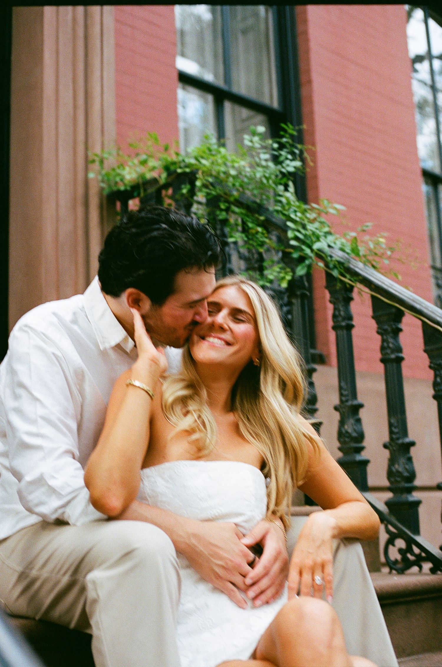 A couple shares a romantic moment on brownstone steps, man kissing woman's cheek as she smiles in a white dress.