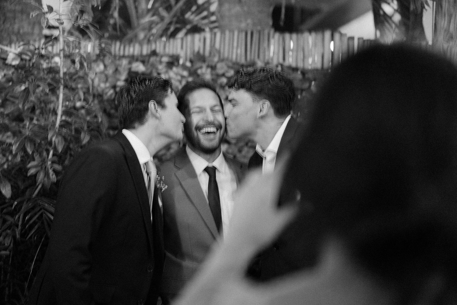 Black and white photo of groom laughing as two friends kiss his cheeks at a wedding celebration.