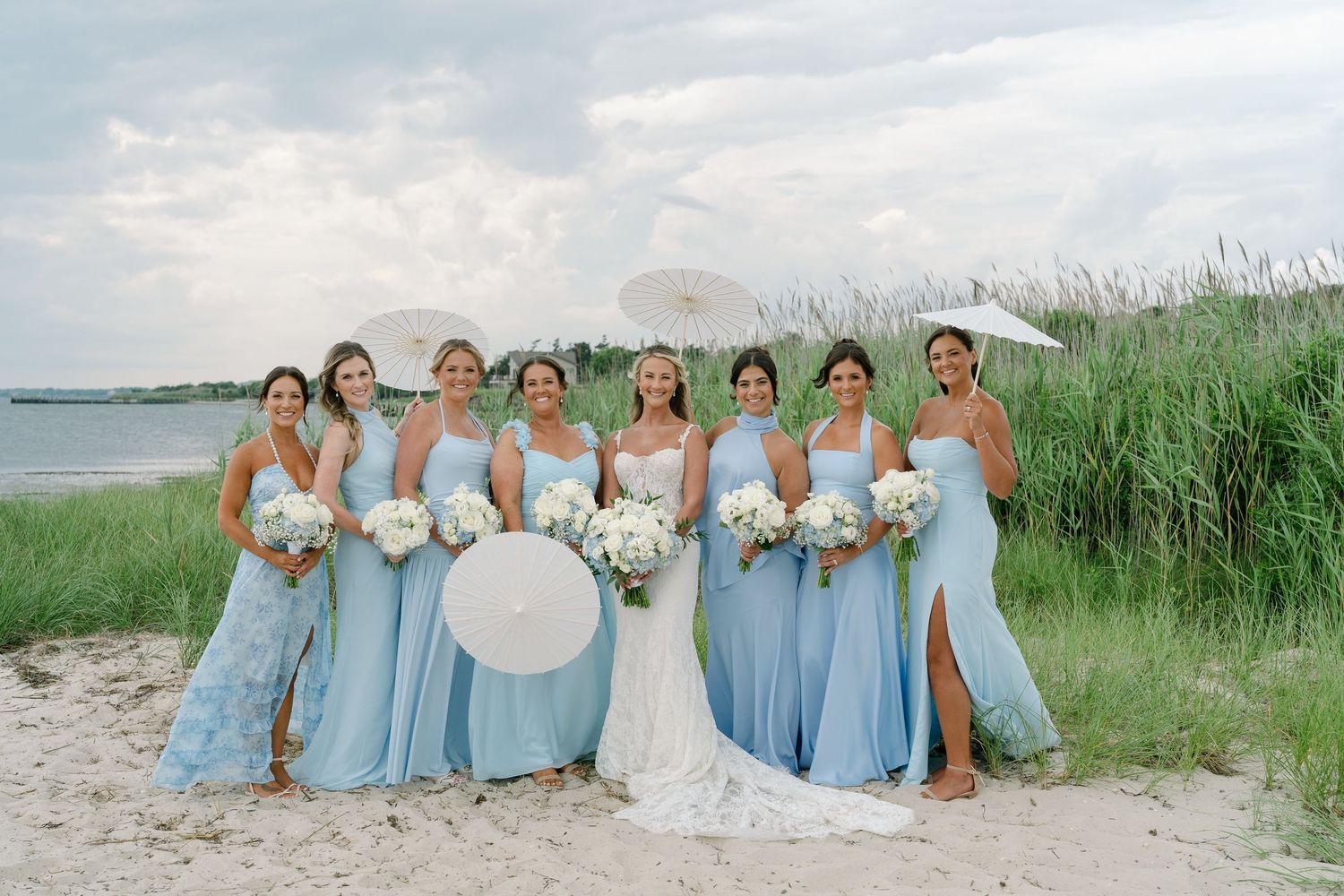 Bride in white gown with bridesmaids in light blue dresses holding white bouquets on a sandy beach.