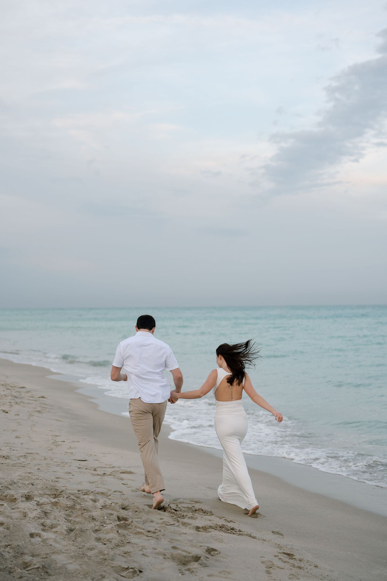 Couple in white clothing walking hand in hand along a serene beach at dusk.