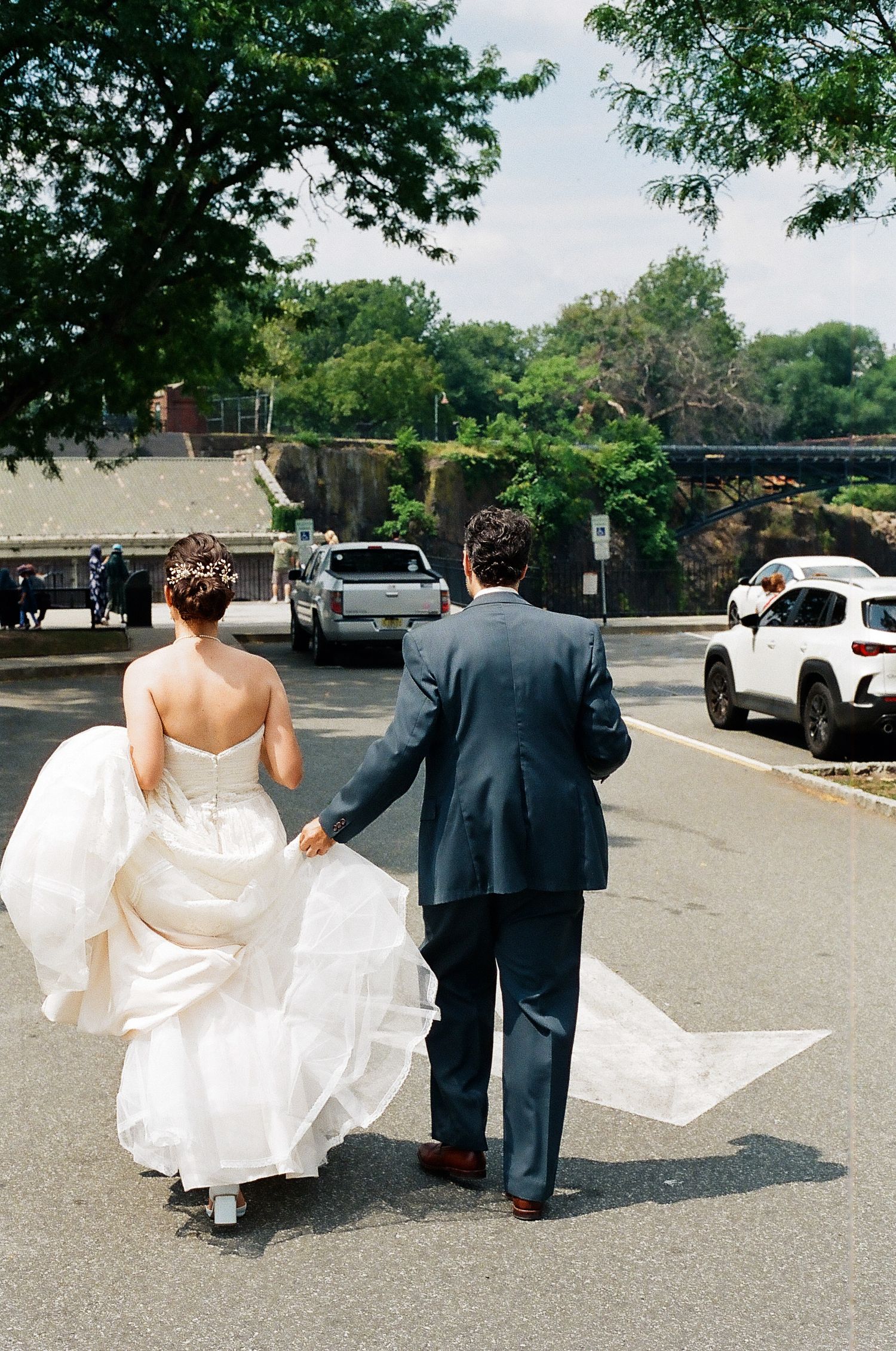 Wedding couple walking across a sunny parking lot while holding the dress train.