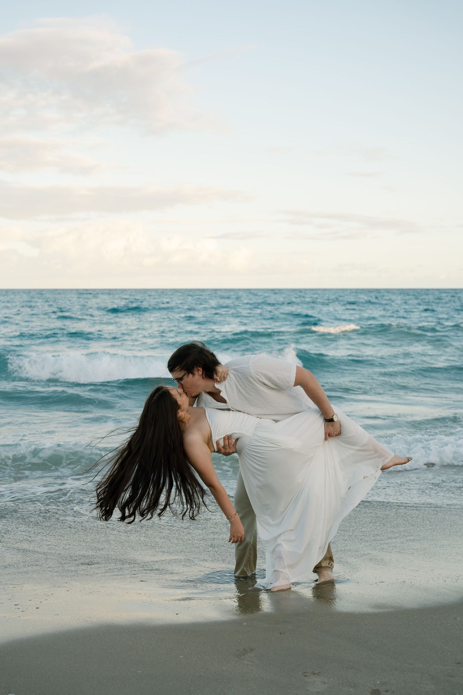 A romantic beach photoshoot features a couple performing a dramatic dip pose by the ocean waves.