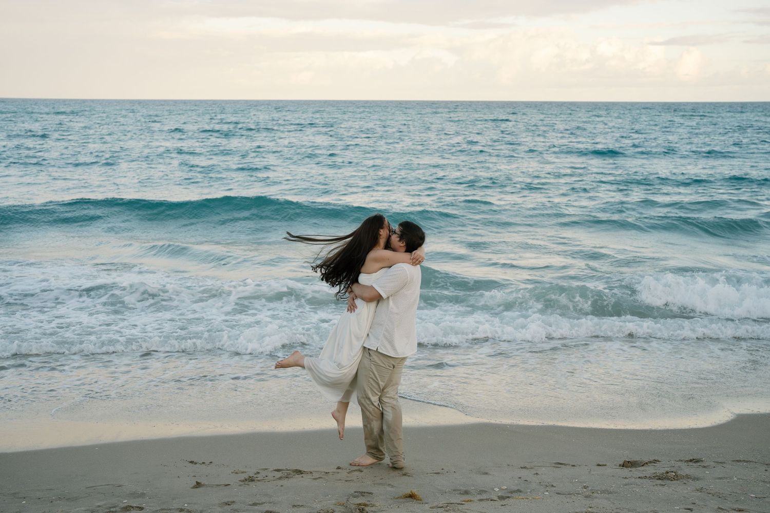 A romantic couple embraces on a beach at sunset with waves crashing in the background and wind blowing through their hair.