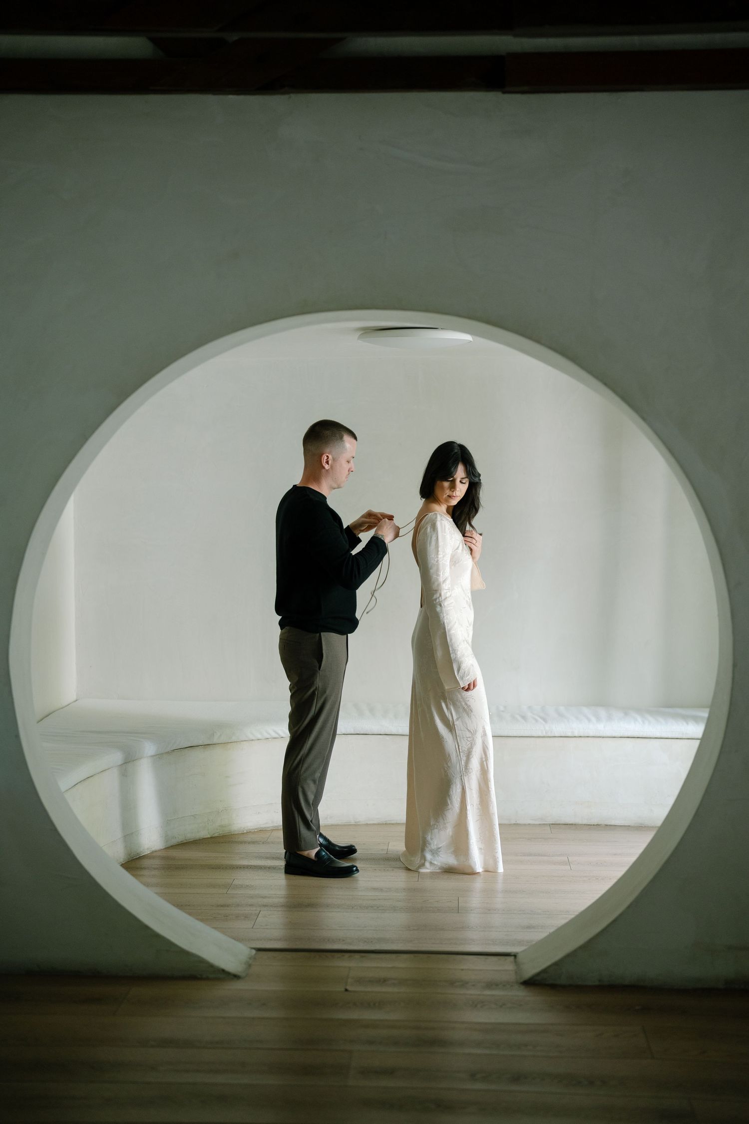 Two figures in contrasting black and white outfits stand in a minimalist curved white room with wooden floors.
