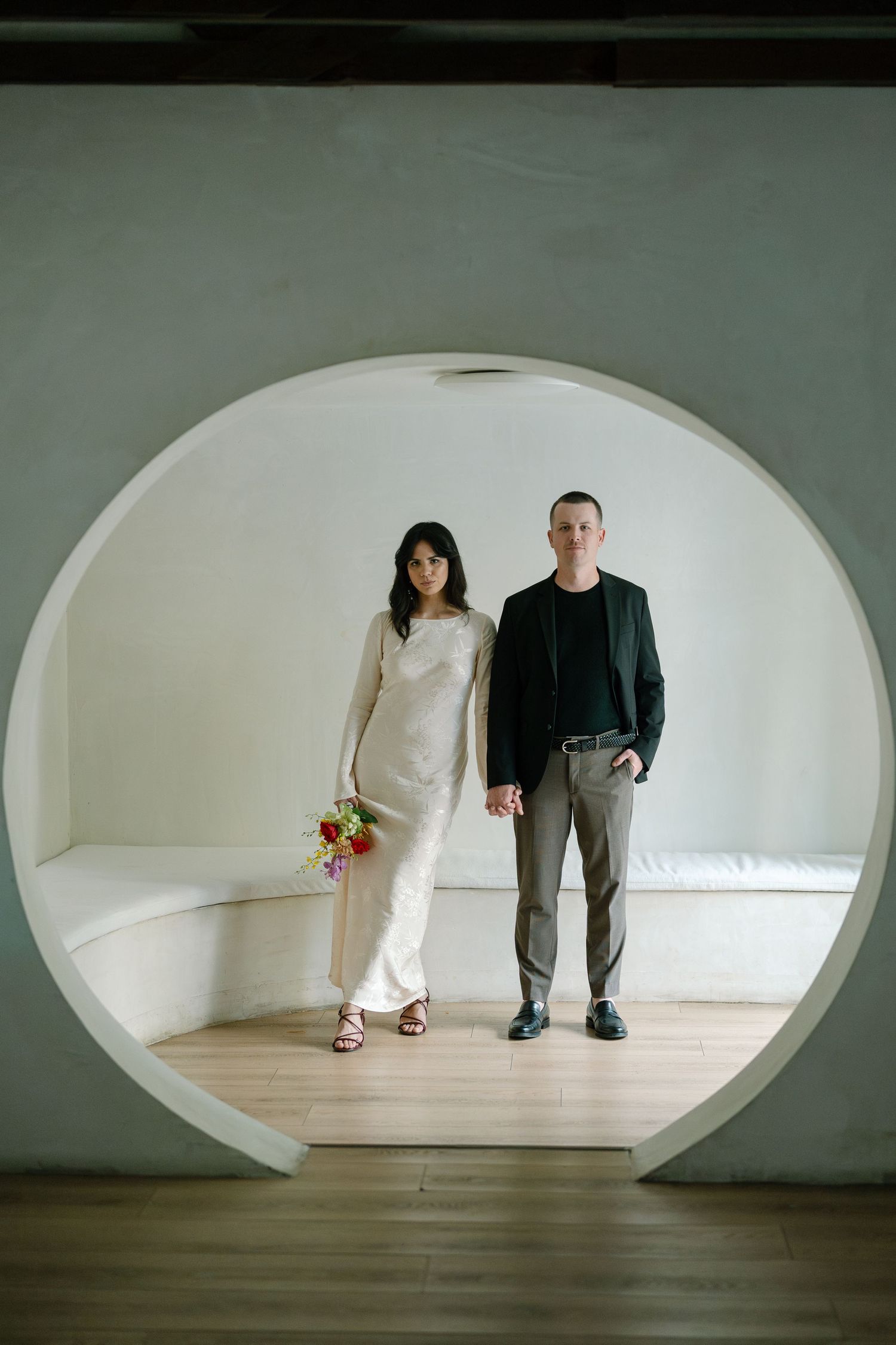 Minimalist photos of two people standing in an oval-framed white room with wooden floors.