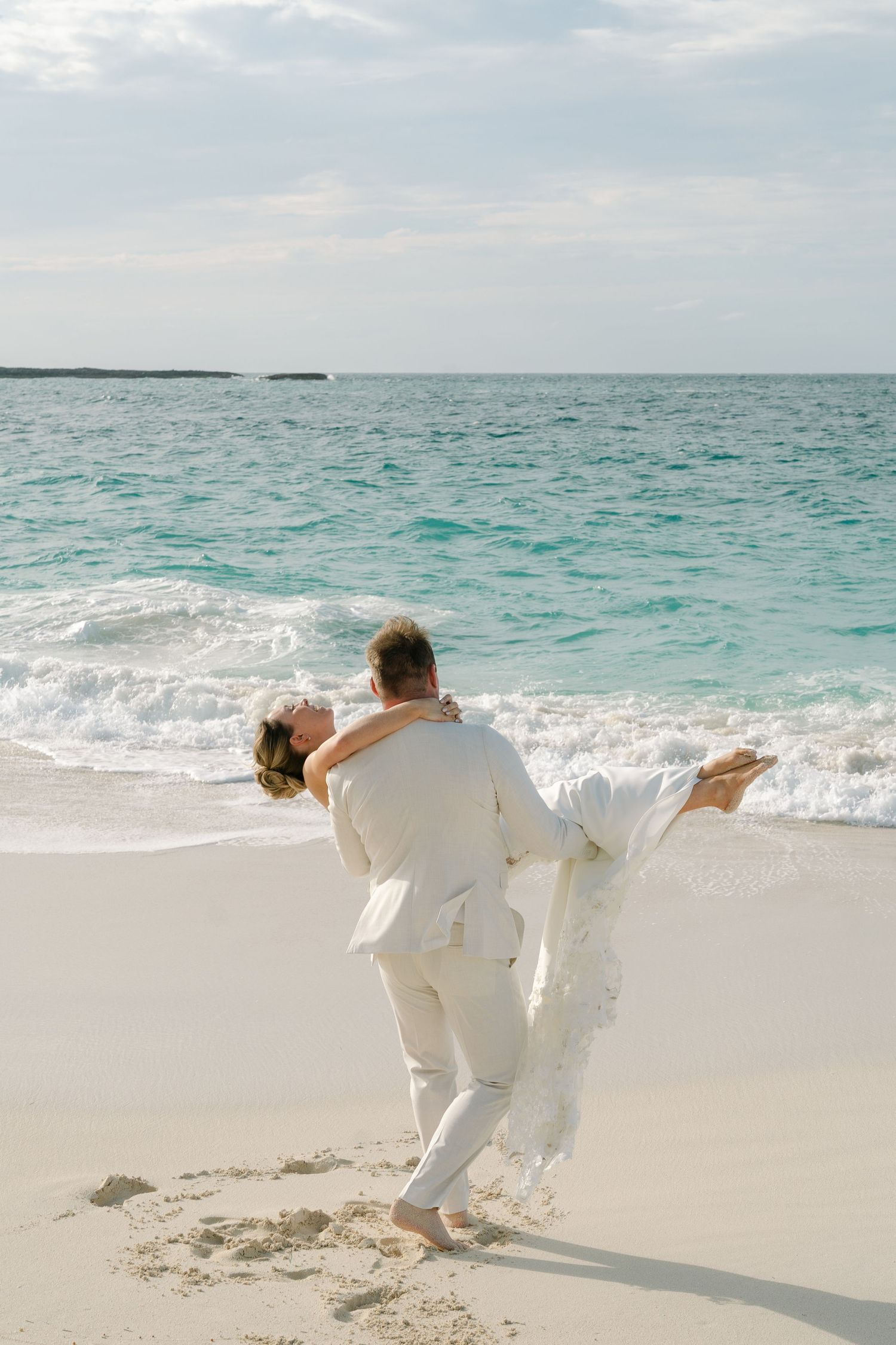 Groom lifting bride on white sand beach with turquoise ocean waves in background.