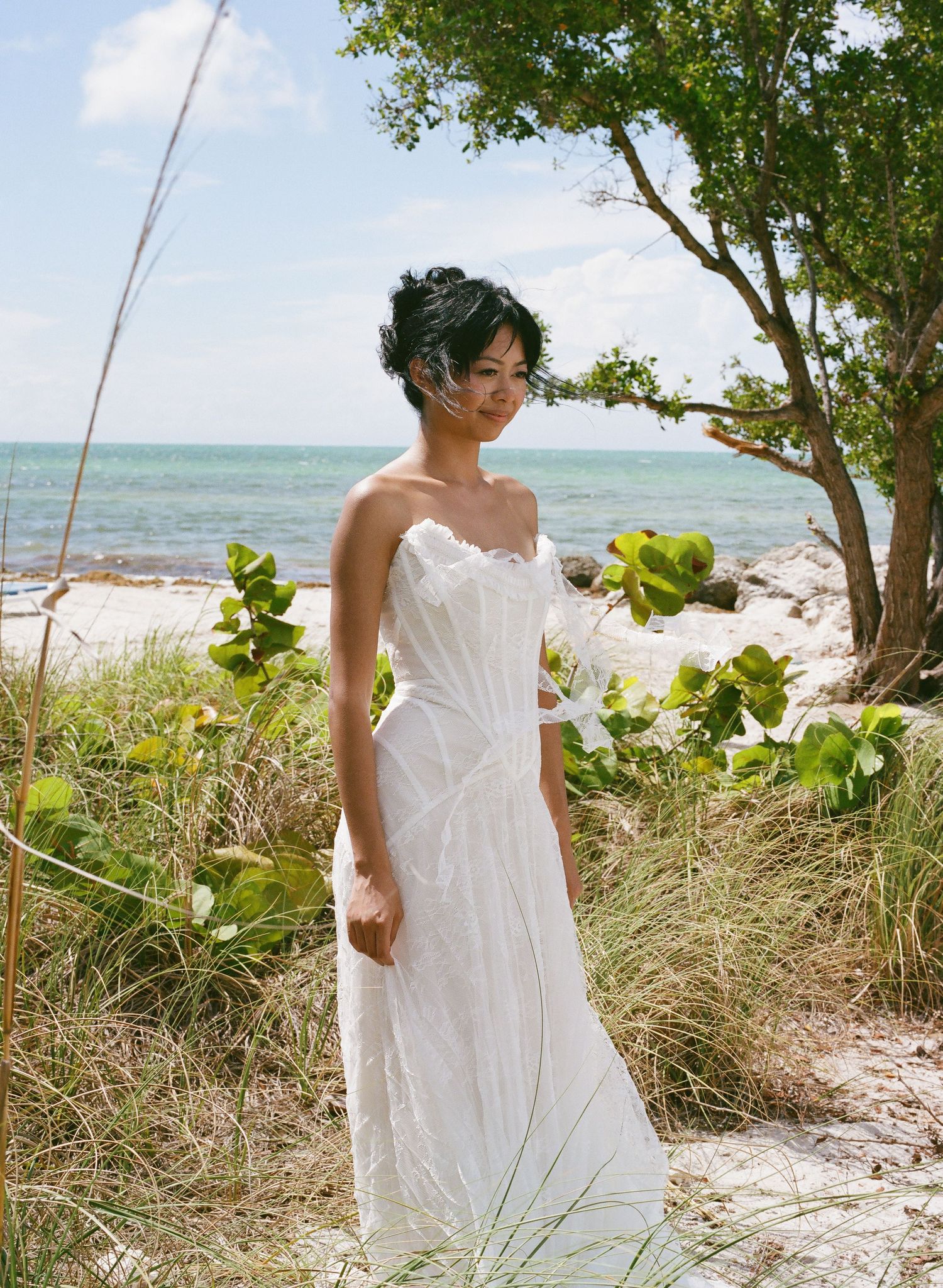 Bride in white strapless gown posing on a tropical beach surrounded by lush greenery and ocean views.