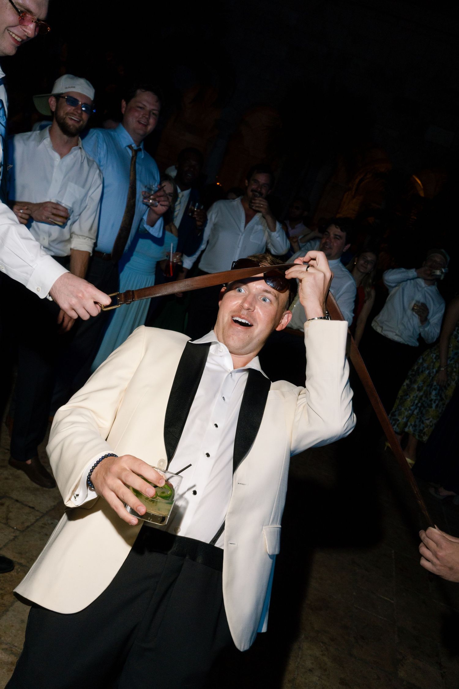 Laughing man in white tuxedo jacket doing the limbo under a stick while holding a cocktail at wedding.
