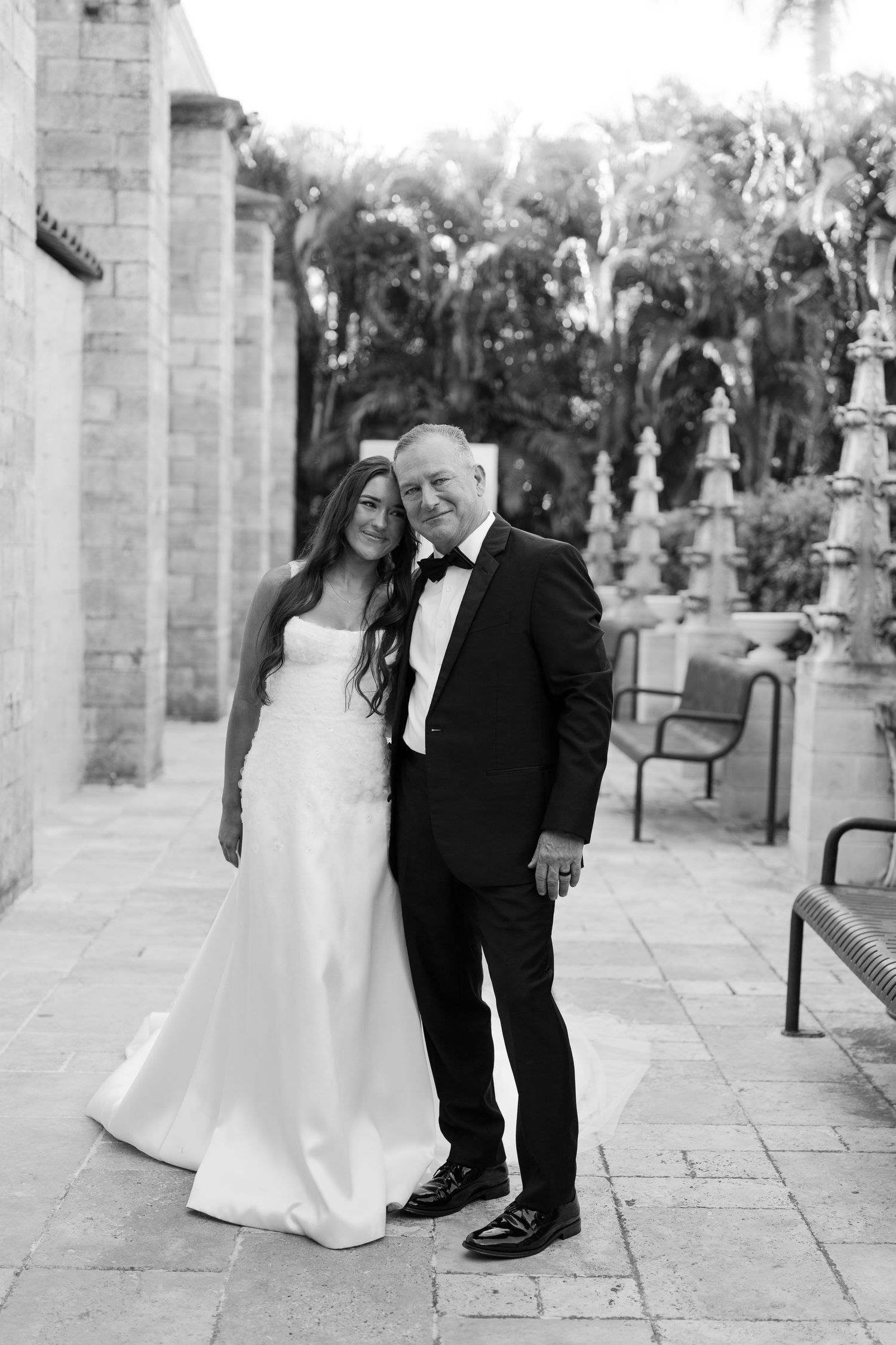 Black and white portrait of bride and older gentleman in tuxedo posing at elegant stone venue.
