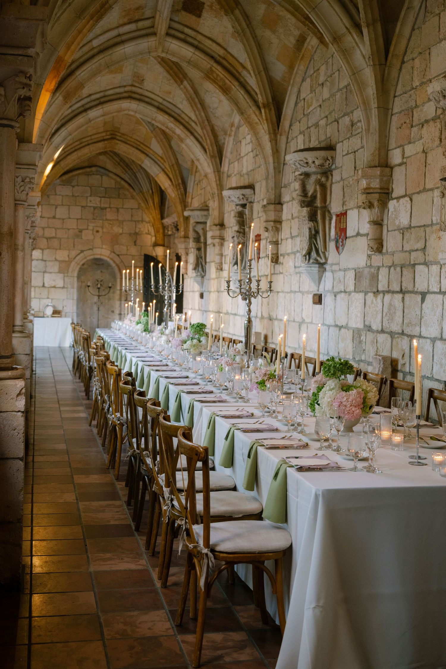 Elegant wedding reception in a historic stone cloister with long candlelit table, green linens, and wooden cross-back chairs.