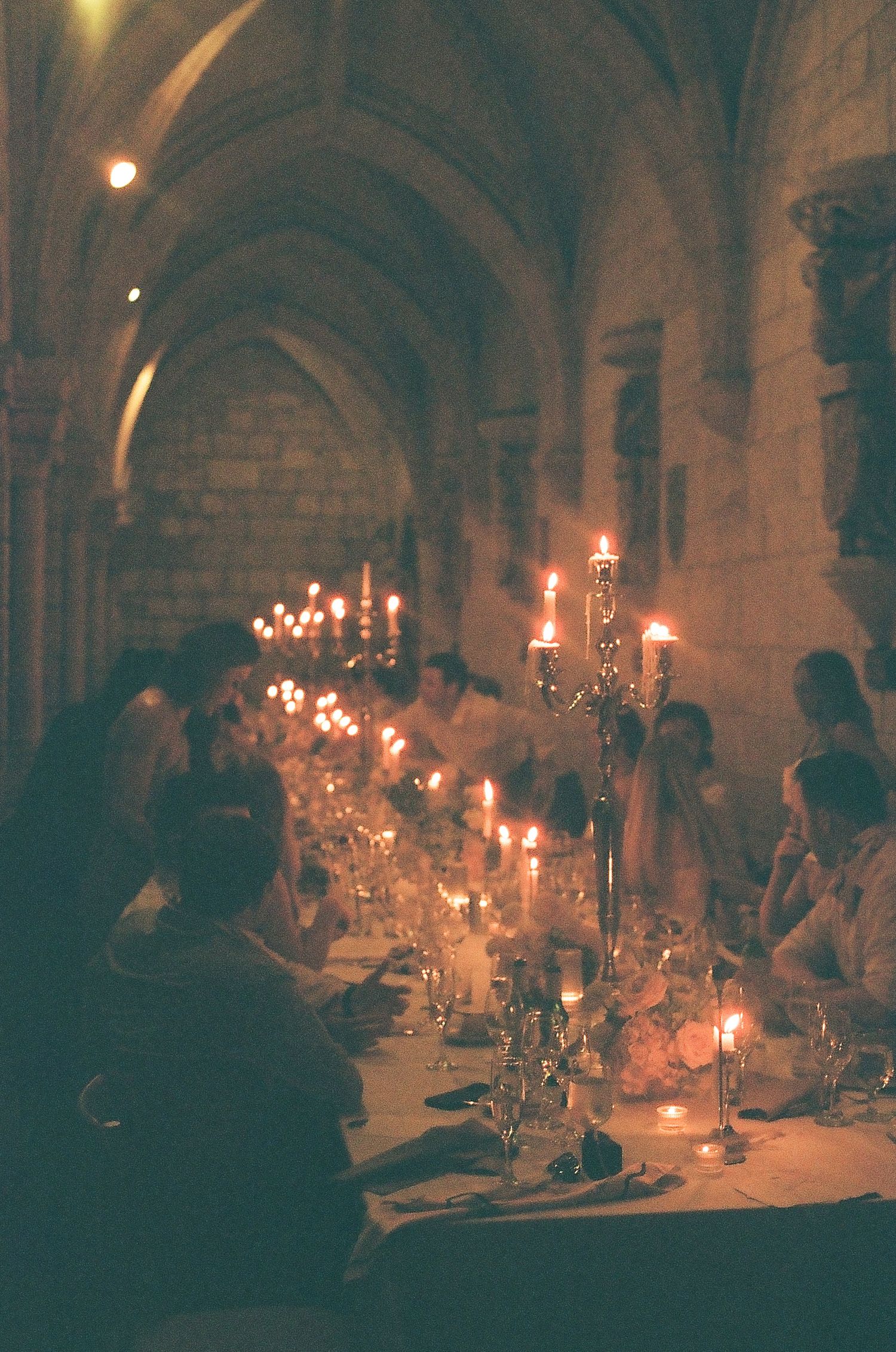 Elegant candlelit dinner in a gothic stone cathedral with long dining table and candelabras glowing warmly.
