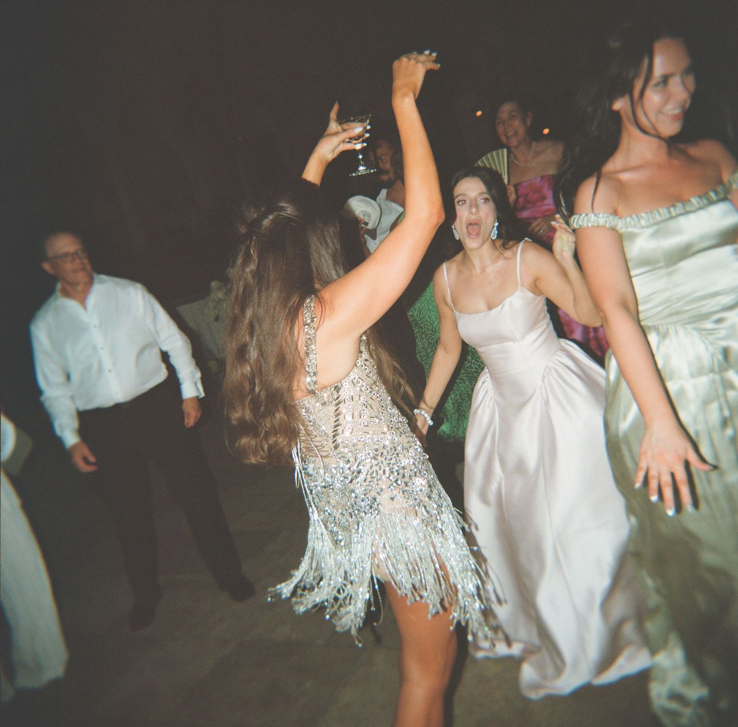 Bride and guests dancing energetically at a wedding reception, woman in sparkly fringe dress raises glass.