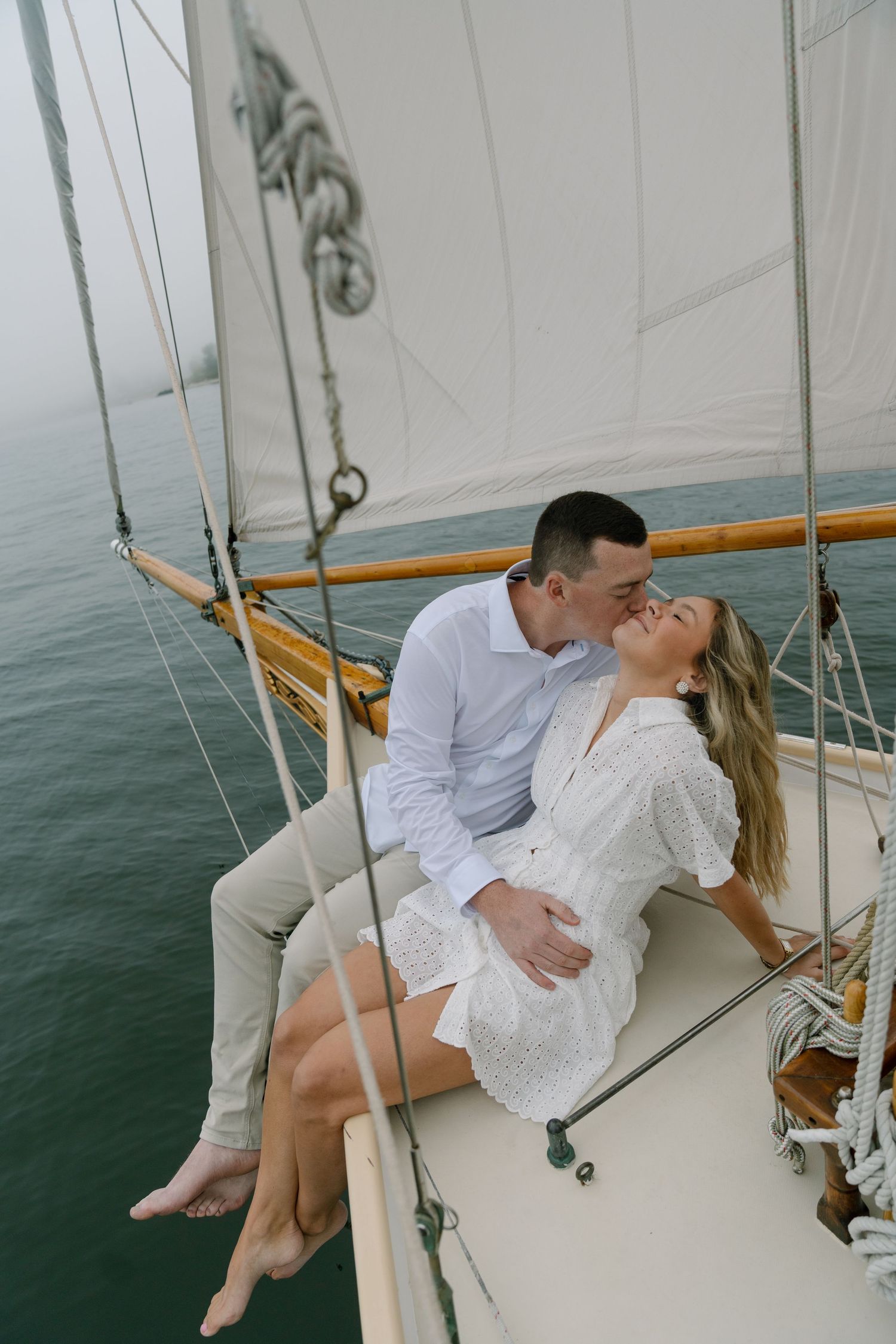 Romantic couple kissing on the bow of a classic sailboat with white sails and turquoise ocean water below.