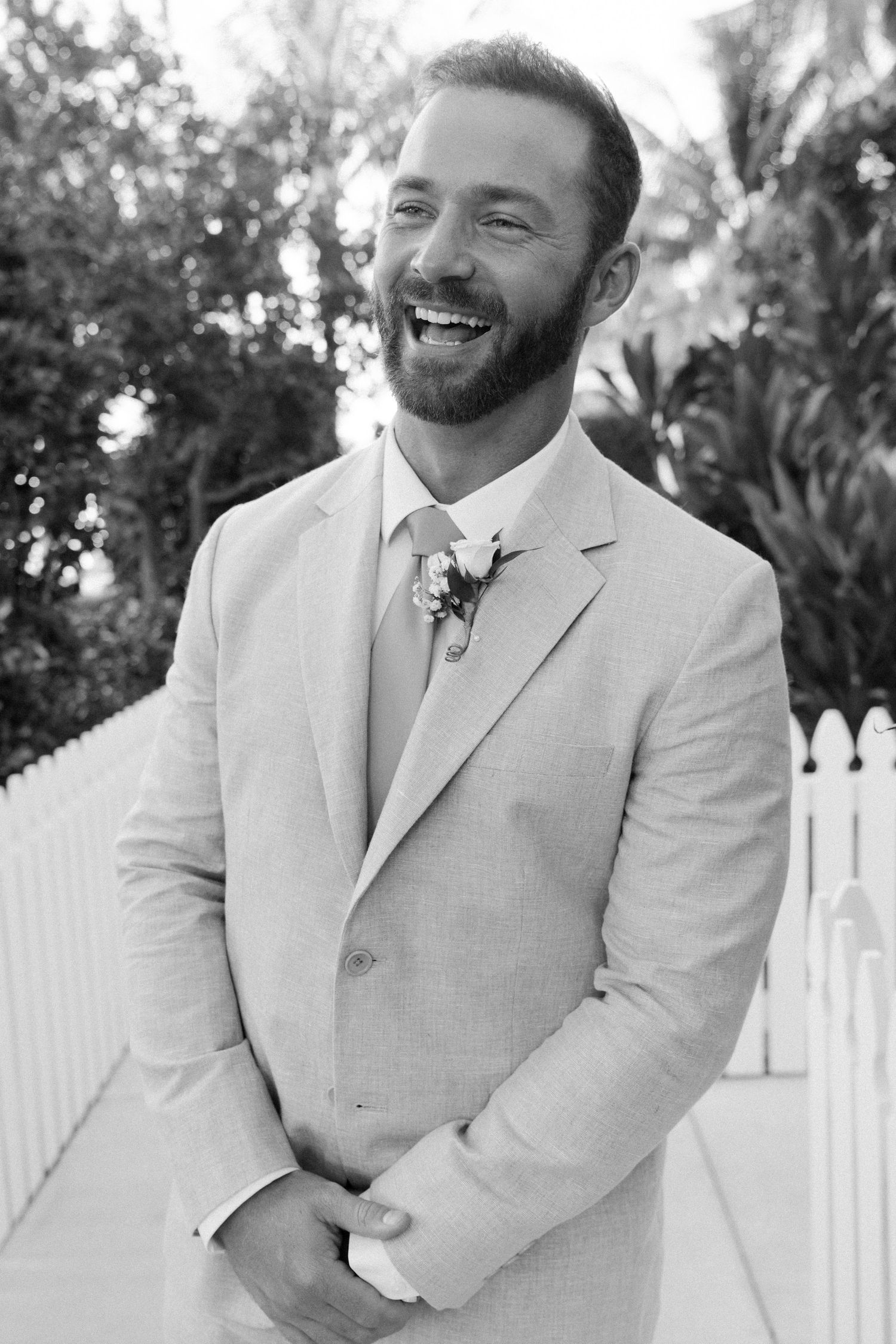 A groom in a light beige suit smiles joyfully in a series of outdoor wedding portraits.