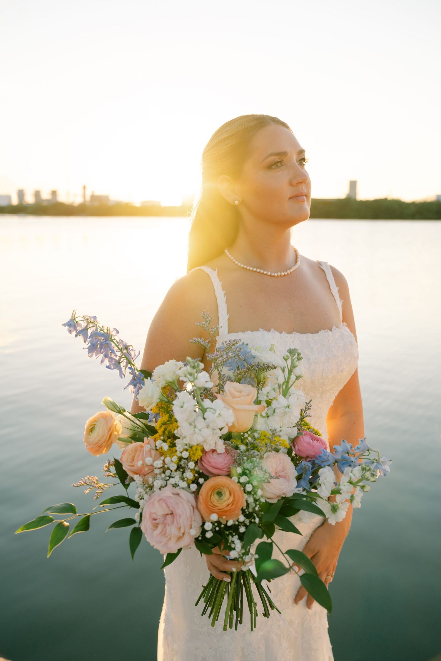 A bride in a white dress holds a beautiful bouquet of pink, peach and white flowers by a lake at sunset.