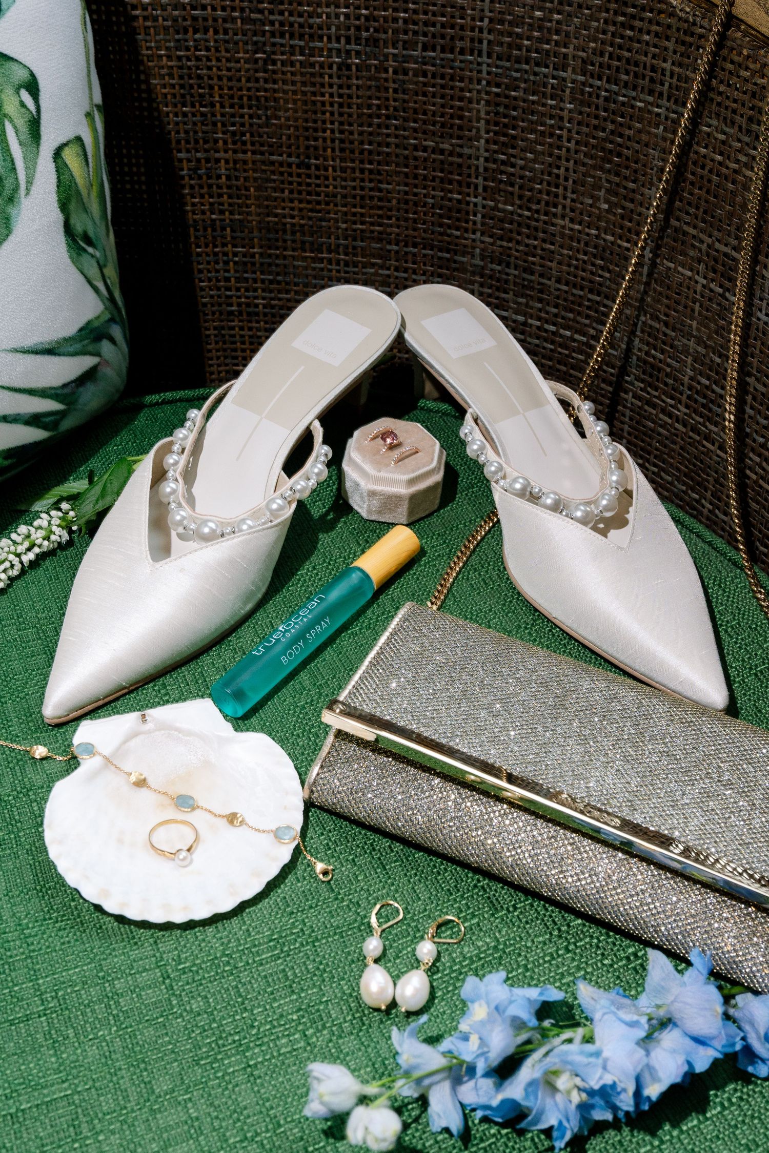 White bridal shoes displayed with blue flowers and jewelry on a green surface.