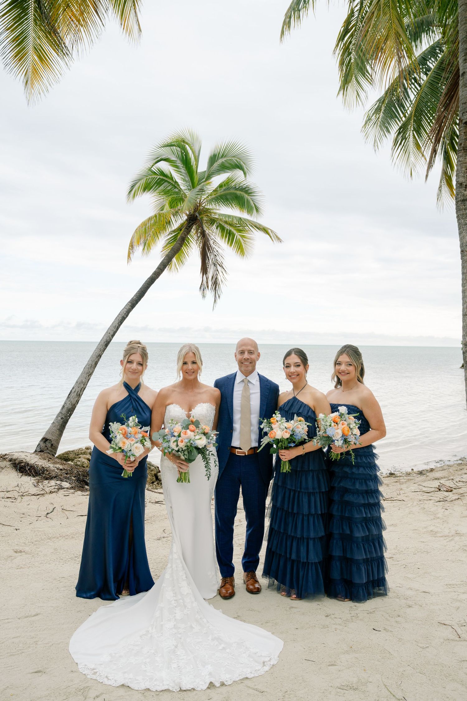 Wedding party in navy blue and white dresses posing on a beach with palm trees.