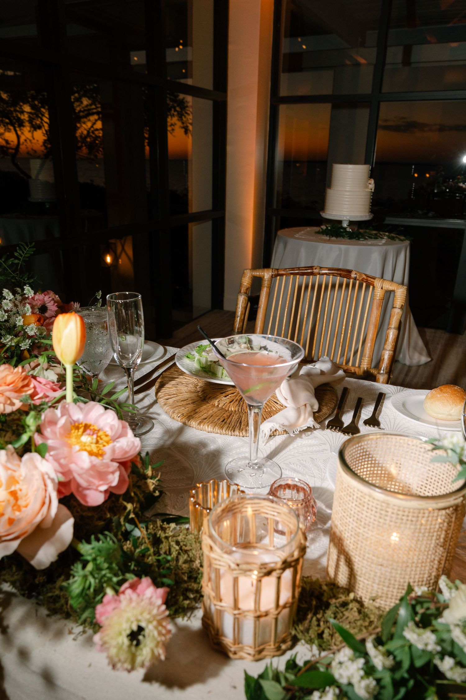 Elegant wedding table setting with pink roses, candlelight and glassware at sunset with a white cake in the background.