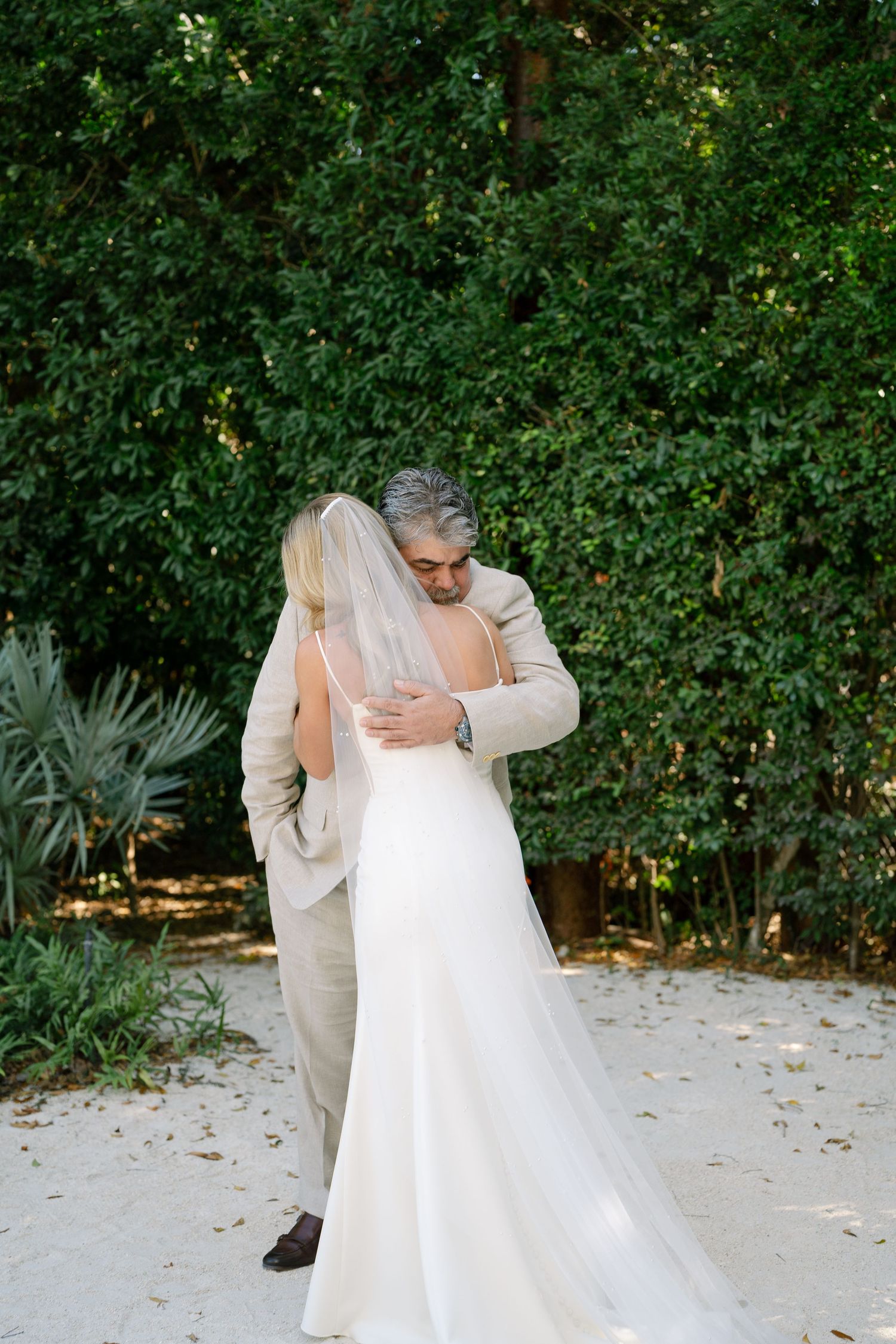 A tender embrace between a bride in white and someone wearing beige captured in an outdoor garden setting.