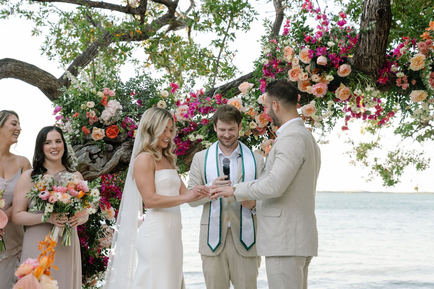 Wedding party gathered under floral-decorated tree arch by the ocean.