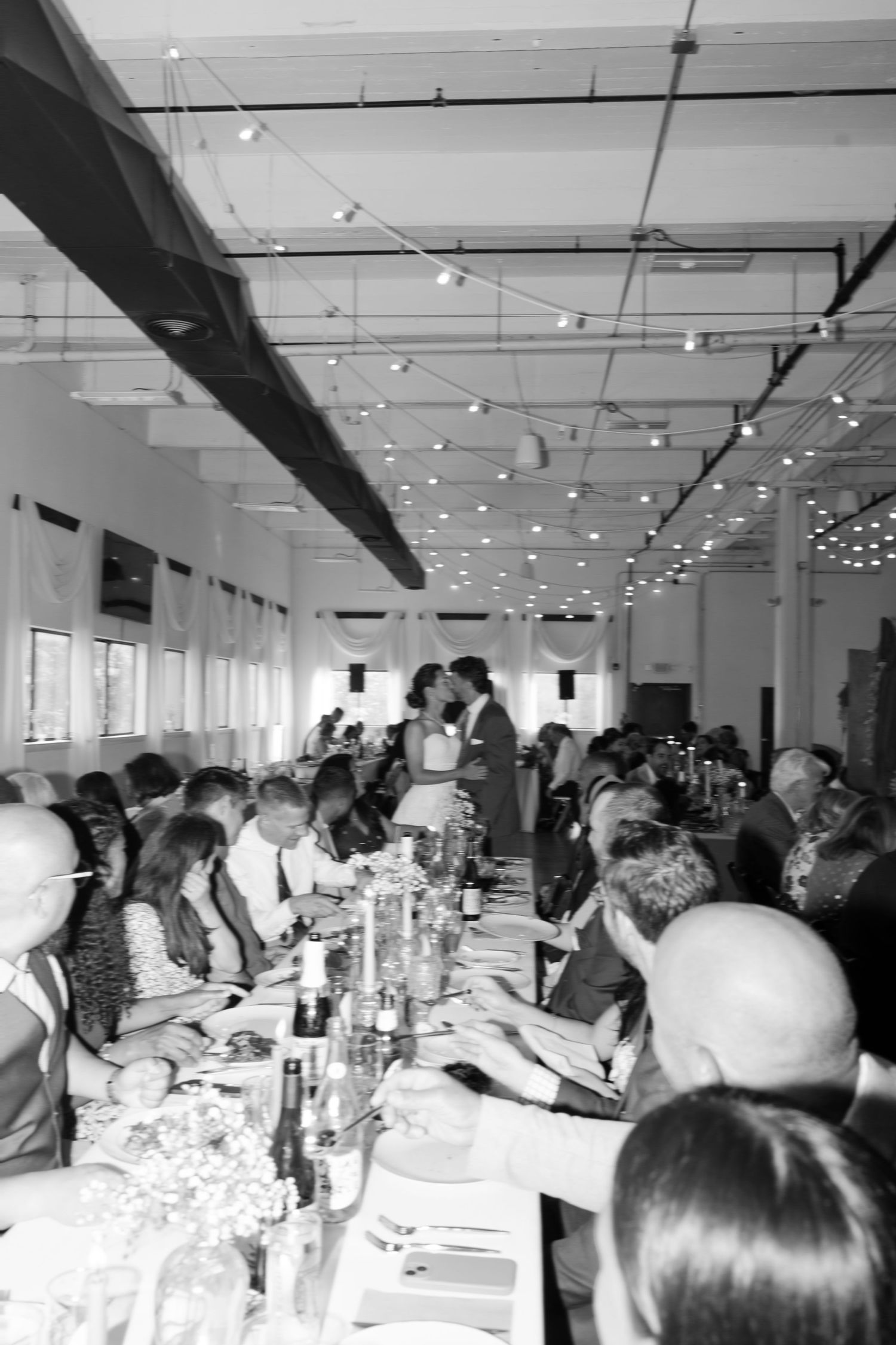 Wedding reception guests seated at long tables in an industrial-style venue with exposed ceiling.