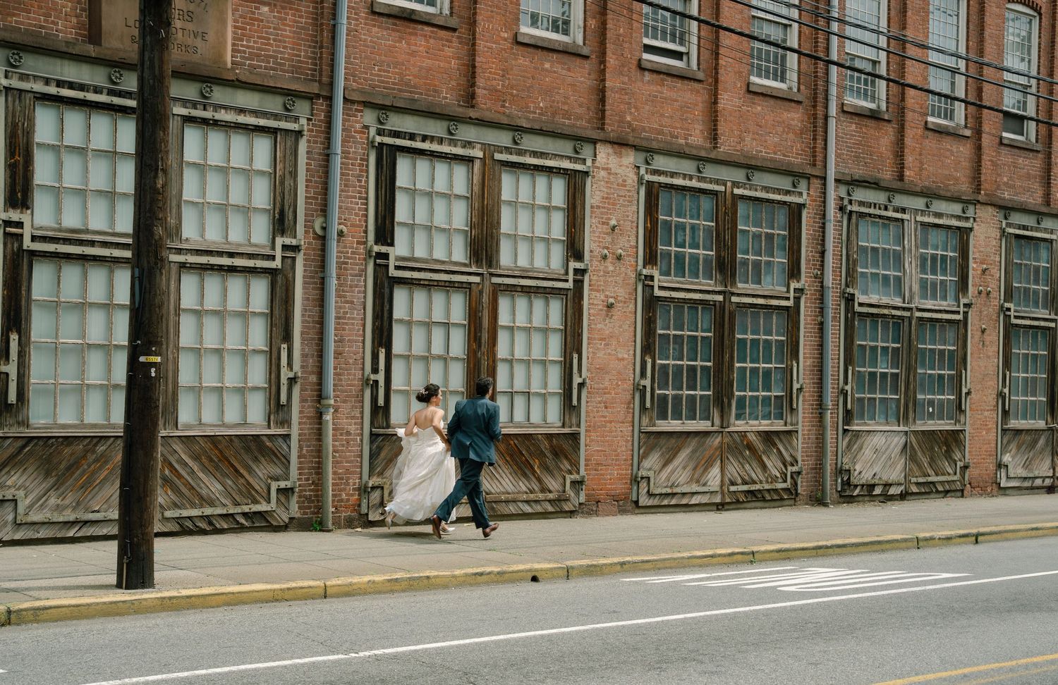 Long brick factory building with industrial windows along a sloped street on an overcast day.