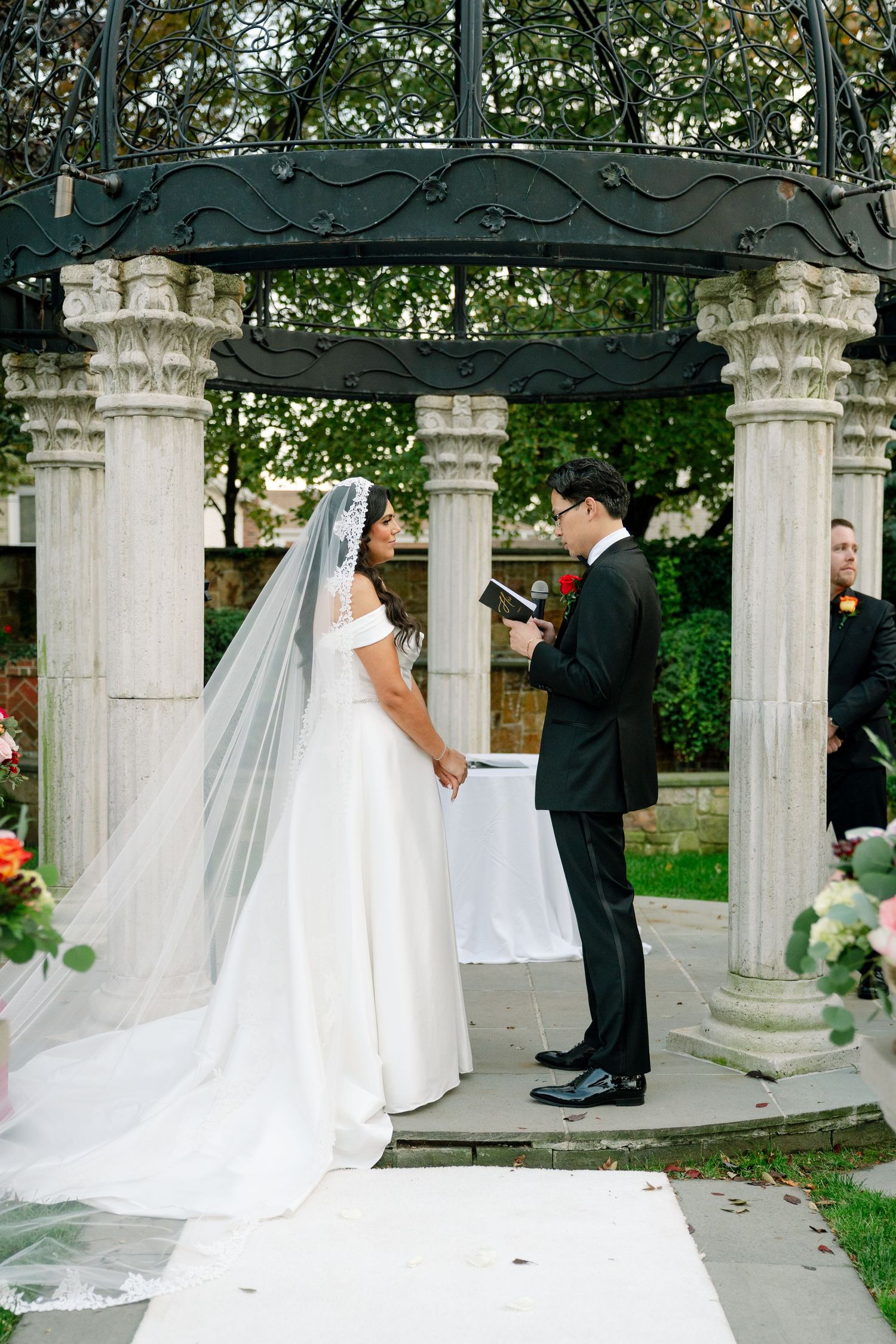 Wedding ceremony under elegant white stone gazebo with flowing bridal veil.
