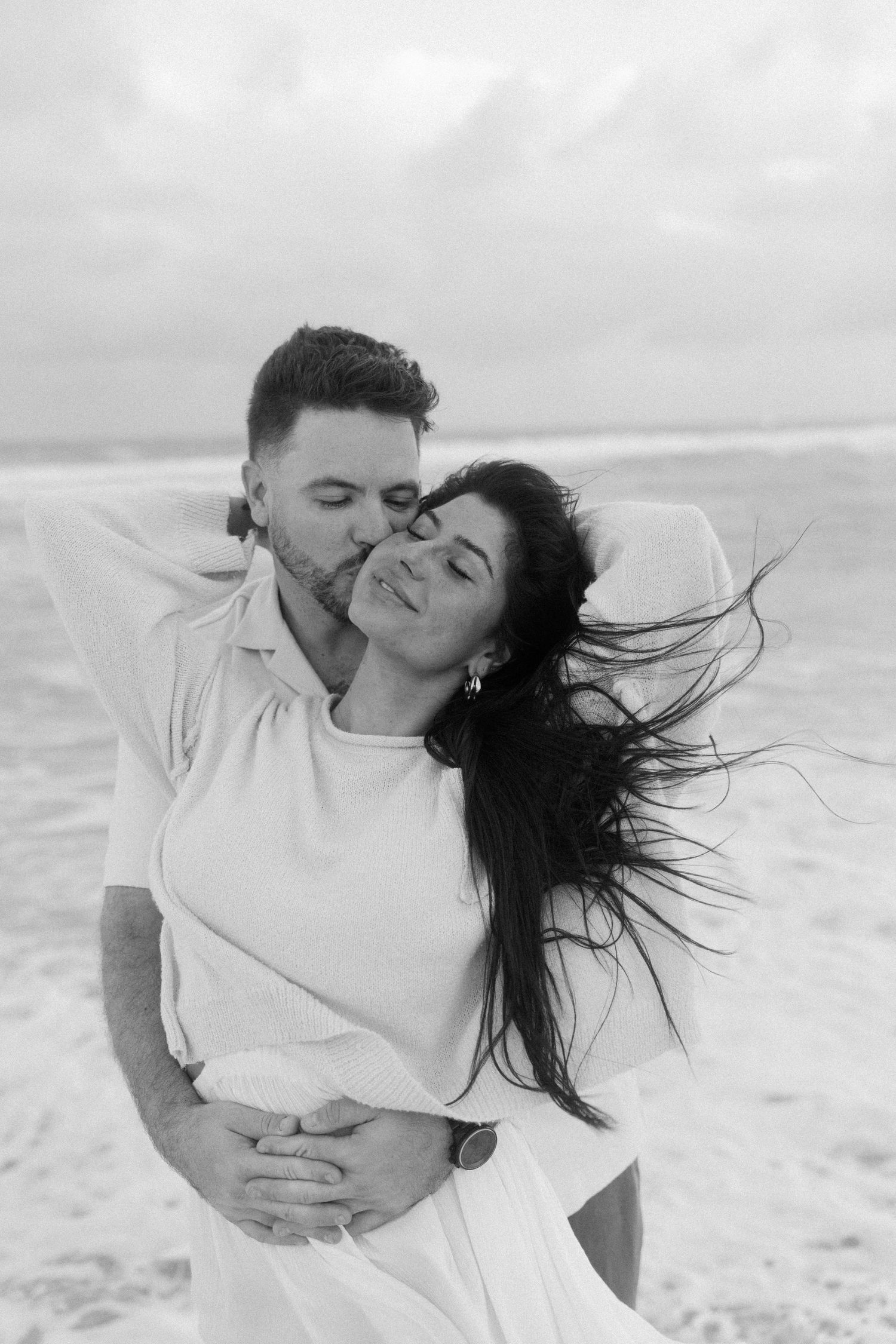 A couple in white clothing shares romantic kisses and embraces on a beach in black and white photography.