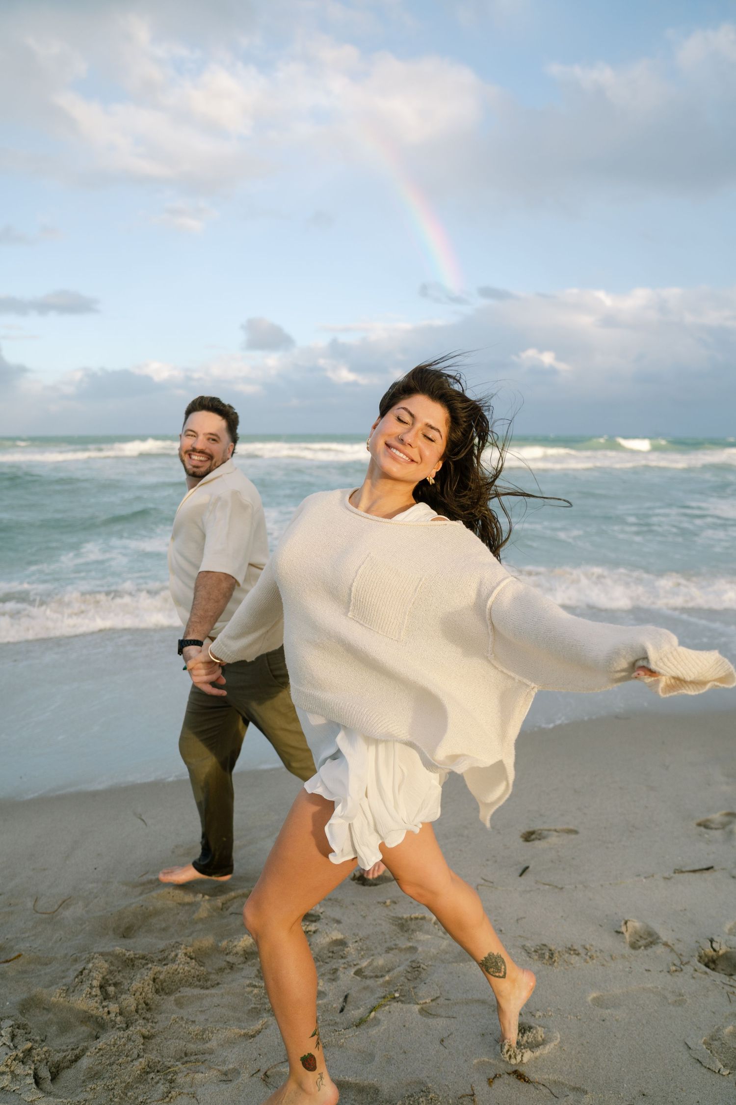 A couple enjoying a playful moment on the beach with crashing waves and a rainbow in the background.