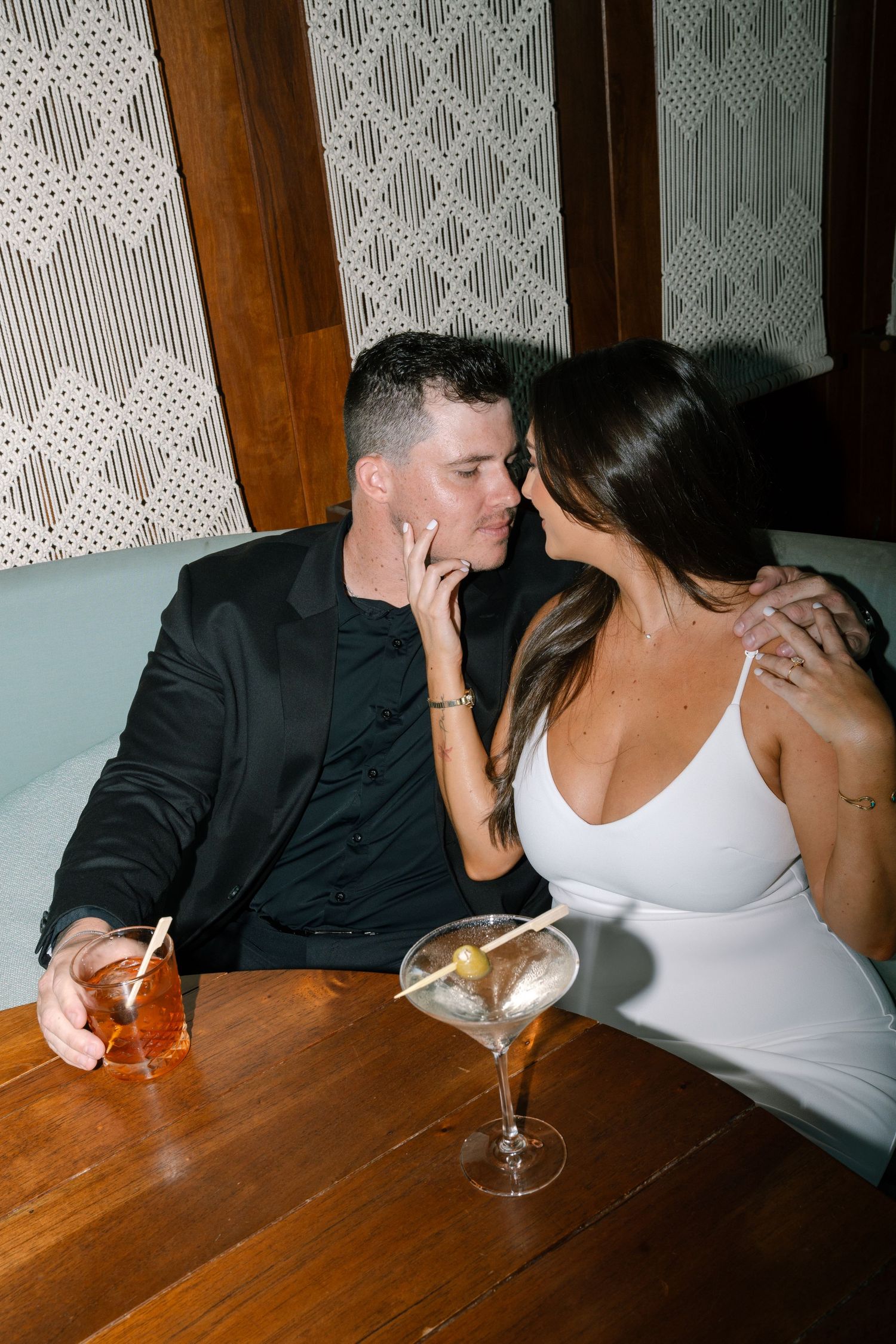 A romantic couple shares an intimate moment at a restaurant table with cocktails and formal attire.