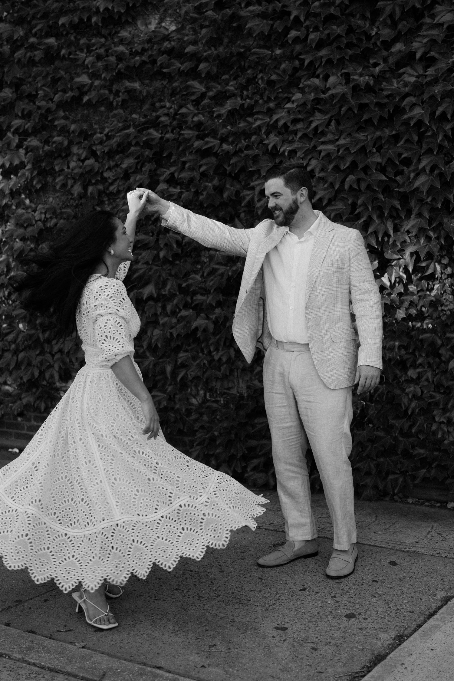 Black and white photo of a couple dancing on a sidewalk, man spinning woman in white lace dress before ivy wall.