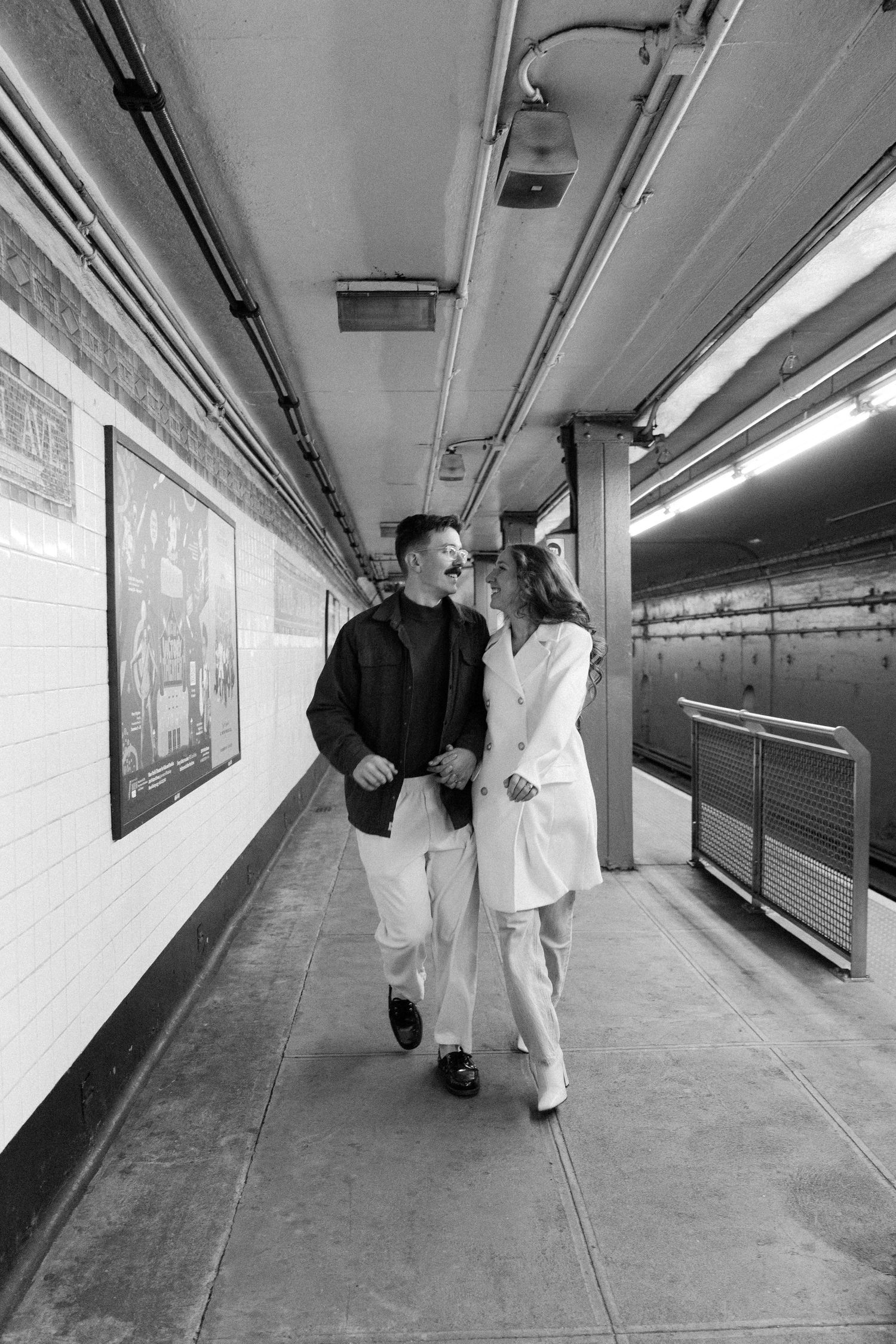 A couple walks together through a stark white subway tunnel corridor.