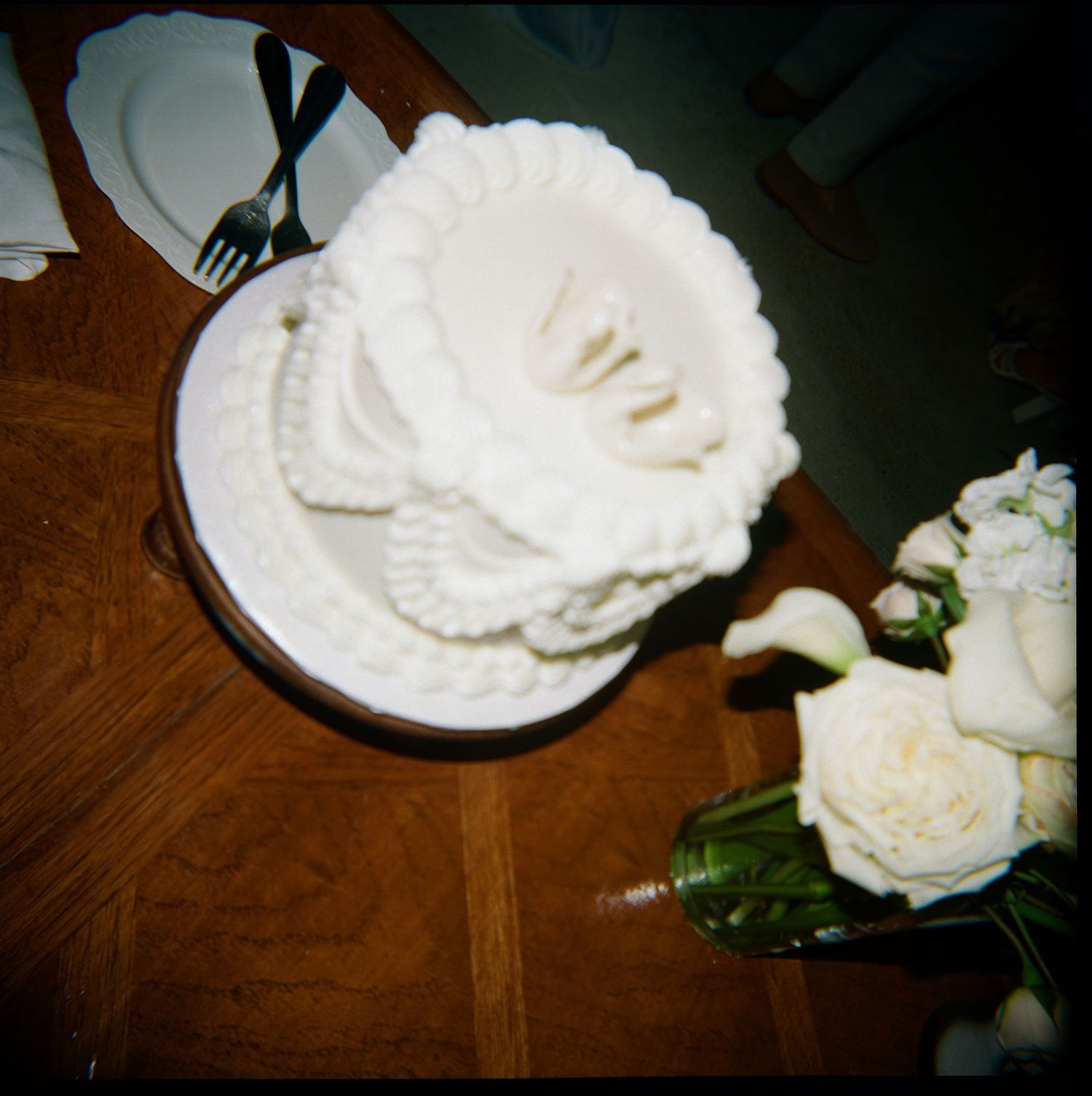 White ruffled wedding cake on plate beside fresh white roses and calla lily bouquet on wooden table.