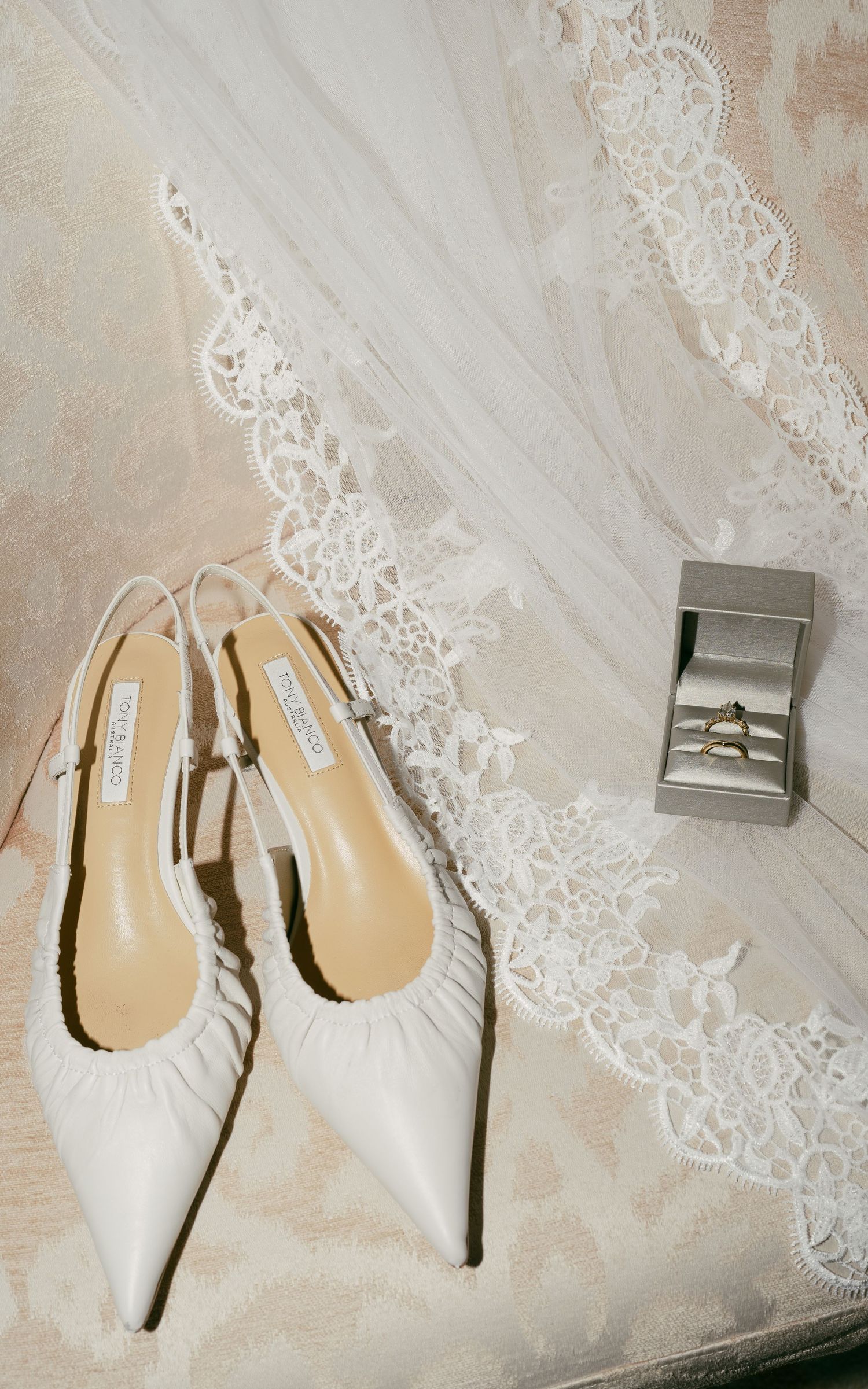 An elegant white bridal setup featuring lace veil details, pointed kitten heel shoes and a silver ring box on cream fabric.