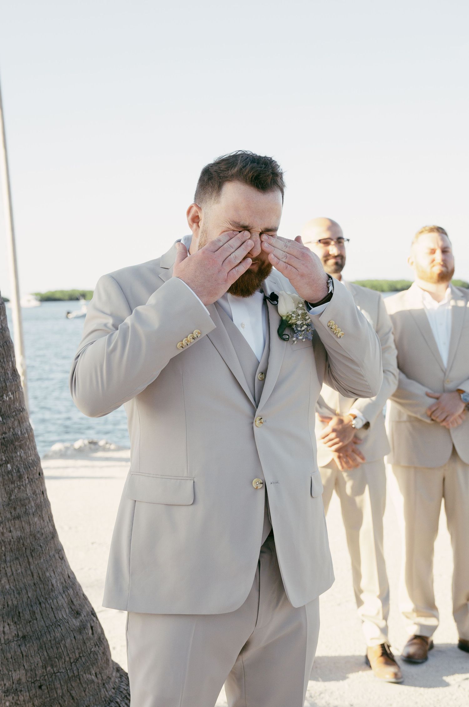 Series of emotional first-look reactions during a beachside wedding ceremony in tan suits near the water.