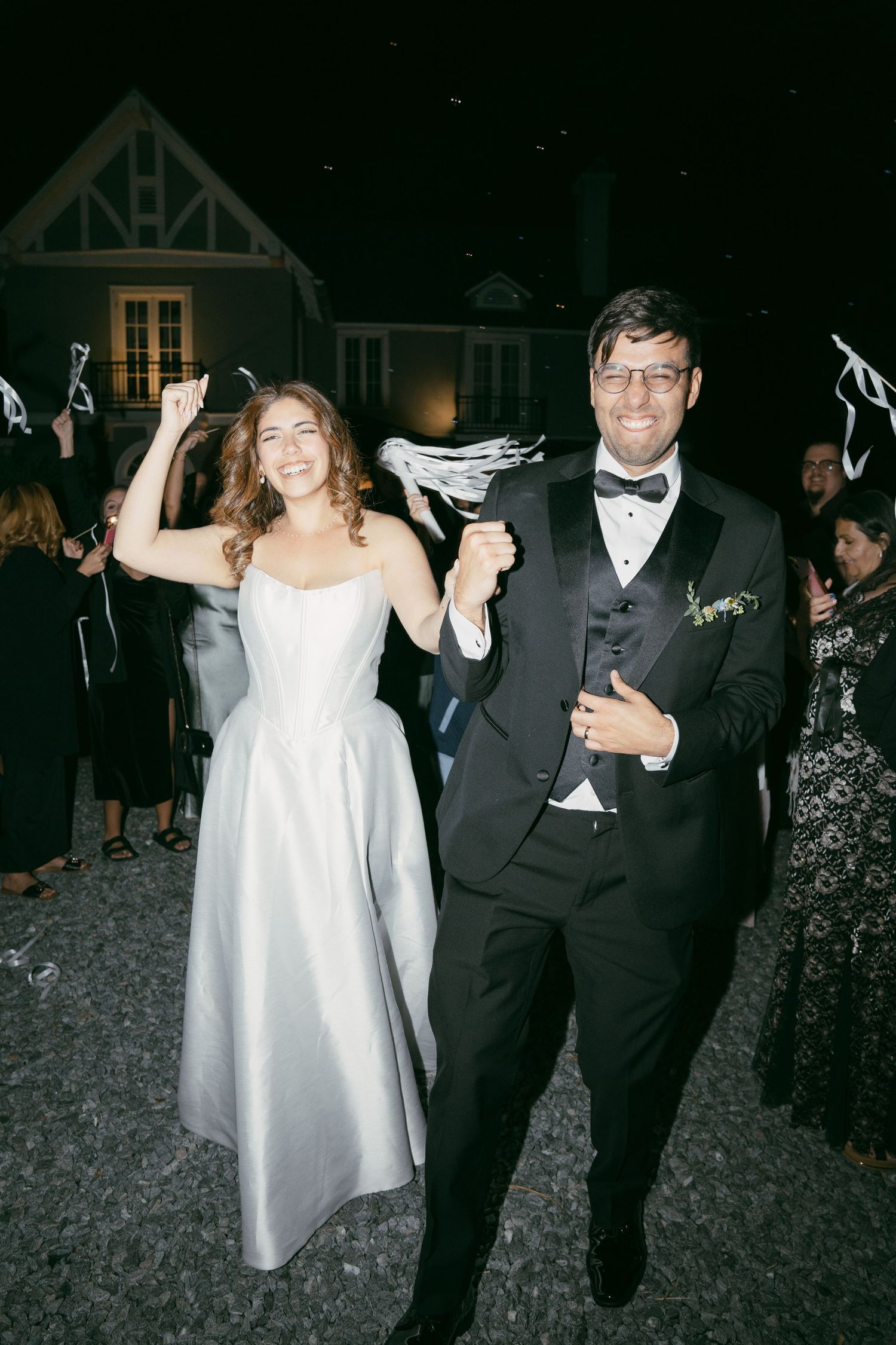 A bride in a white dress and groom in a black tuxedo celebrating while holding shoes at a nighttime wedding reception.