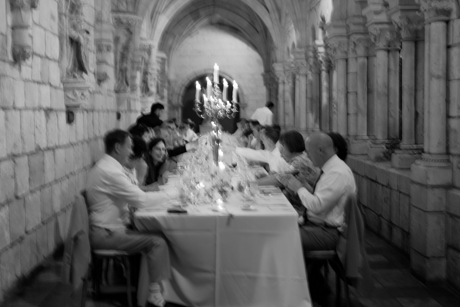 Black and white photo of wedding guests dining at long table in stone cathedral corridor.