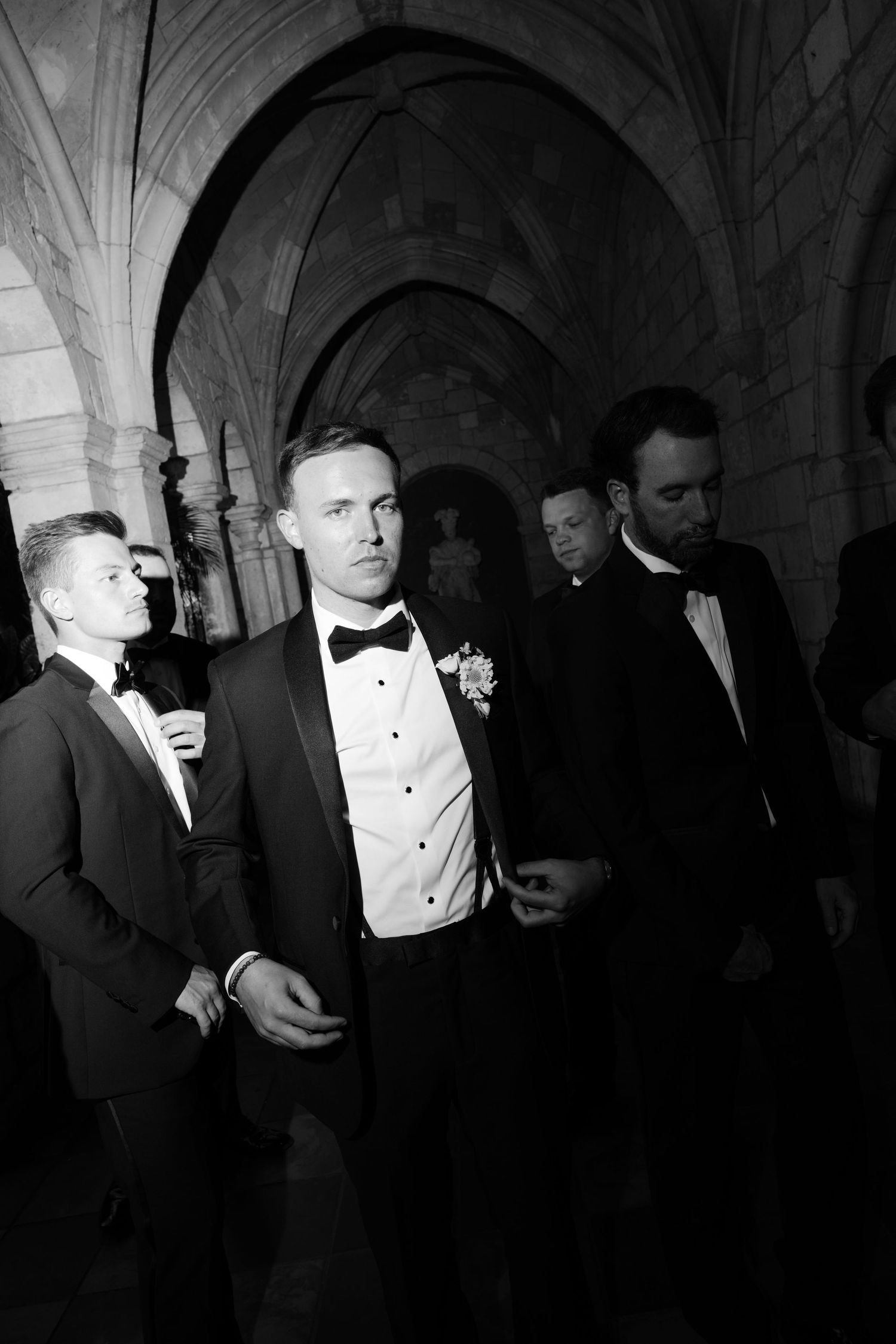 Groomsmen in black tuxedos walk through stone arched corridor at wedding venue in black and white photo.