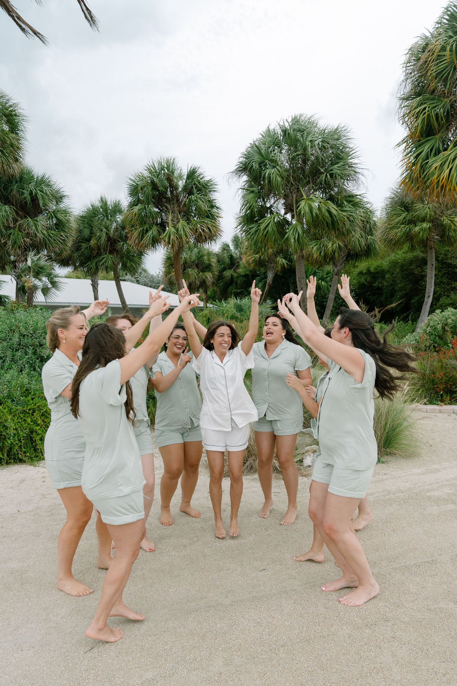 Bride in white pajamas celebrated by bridesmaids in sage green matching sets on sandy beach with palm trees.