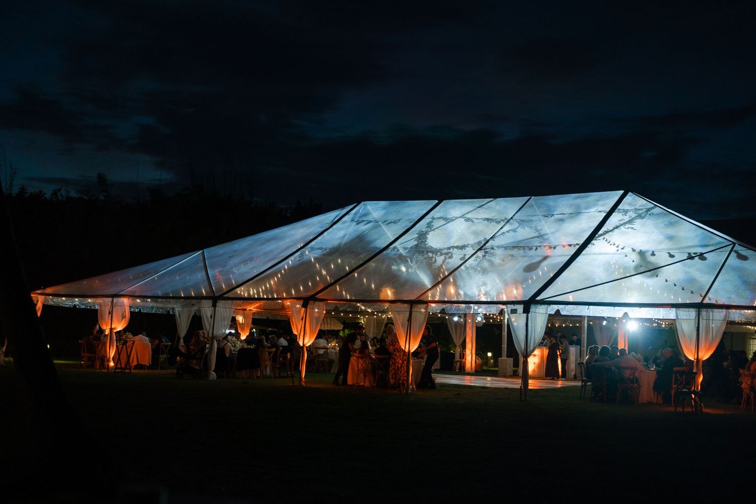 Elegant clear wedding tent glowing with warm lights against a dramatic night sky.