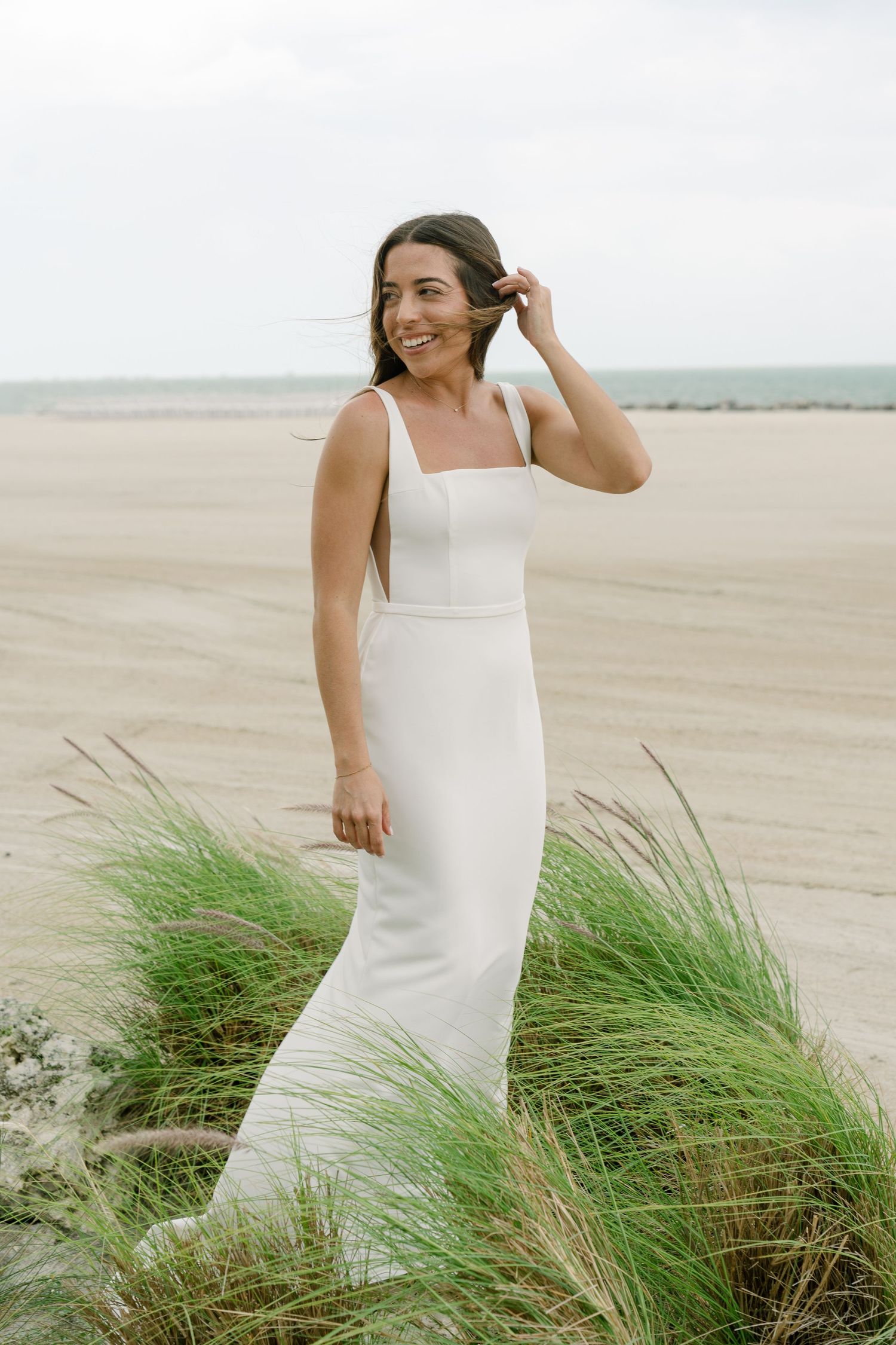 Bride in sleek white square-neck wedding gown smiling on sandy beach with coastal grass.