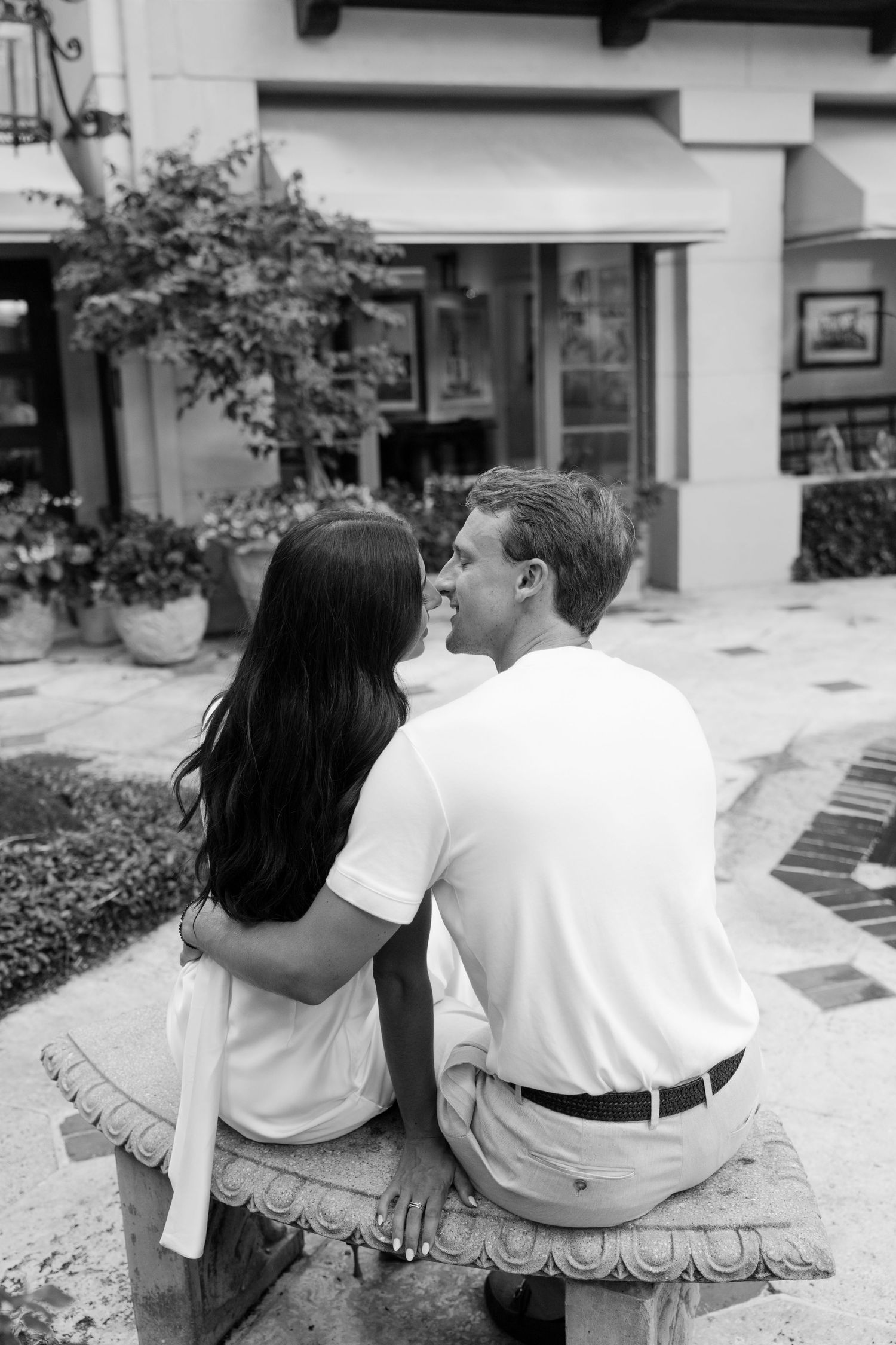 Black and white photo of a couple sitting on a bench, embracing and smiling at each other outside a shop.