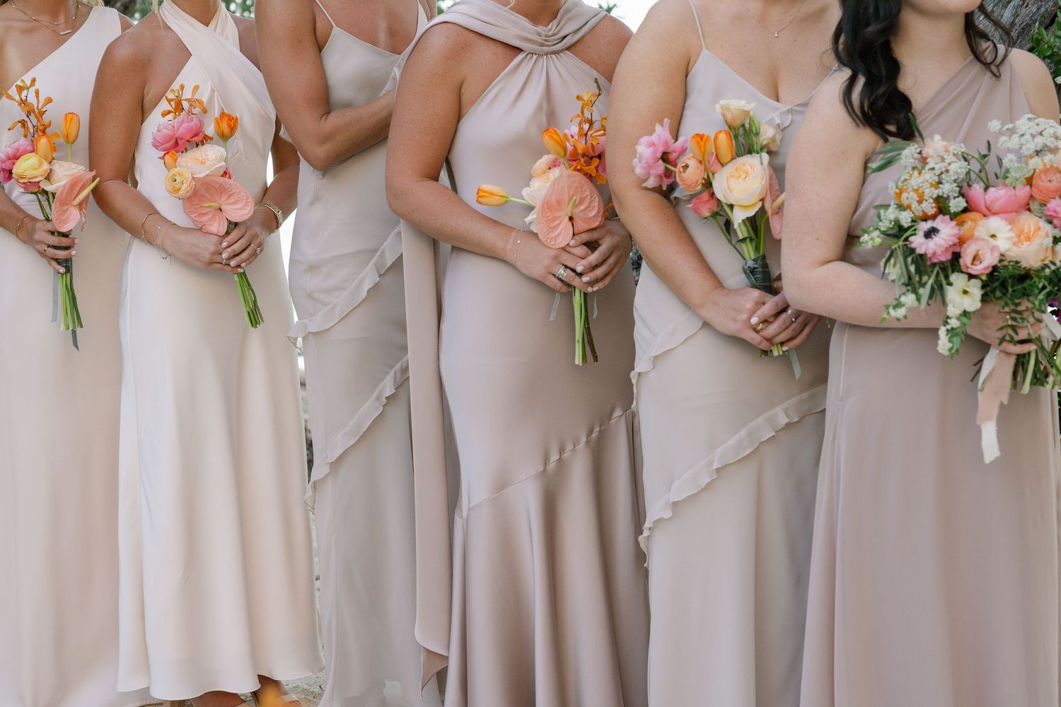 Bridesmaids in neutral-colored dresses holding pink and peach floral bouquets.