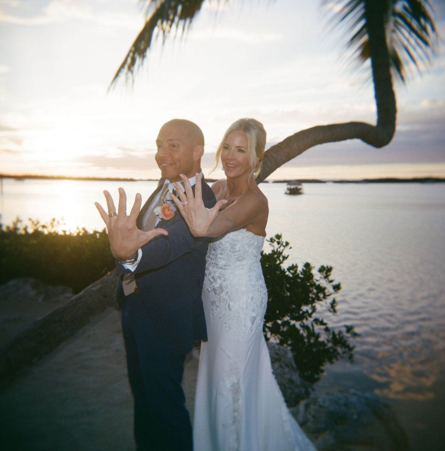 A couple in formal attire dances and poses playfully at sunset by a waterfront location.