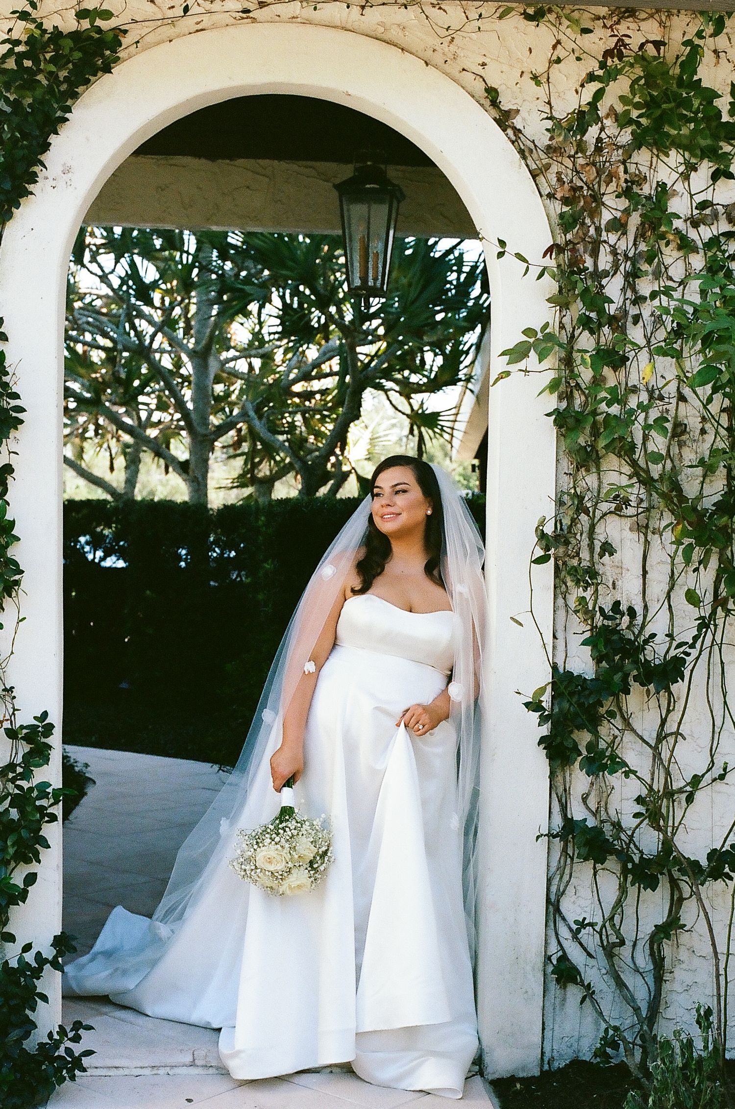 A bride in a white wedding dress poses gracefully under a vine-covered garden pergola in a series of elegant portraits.