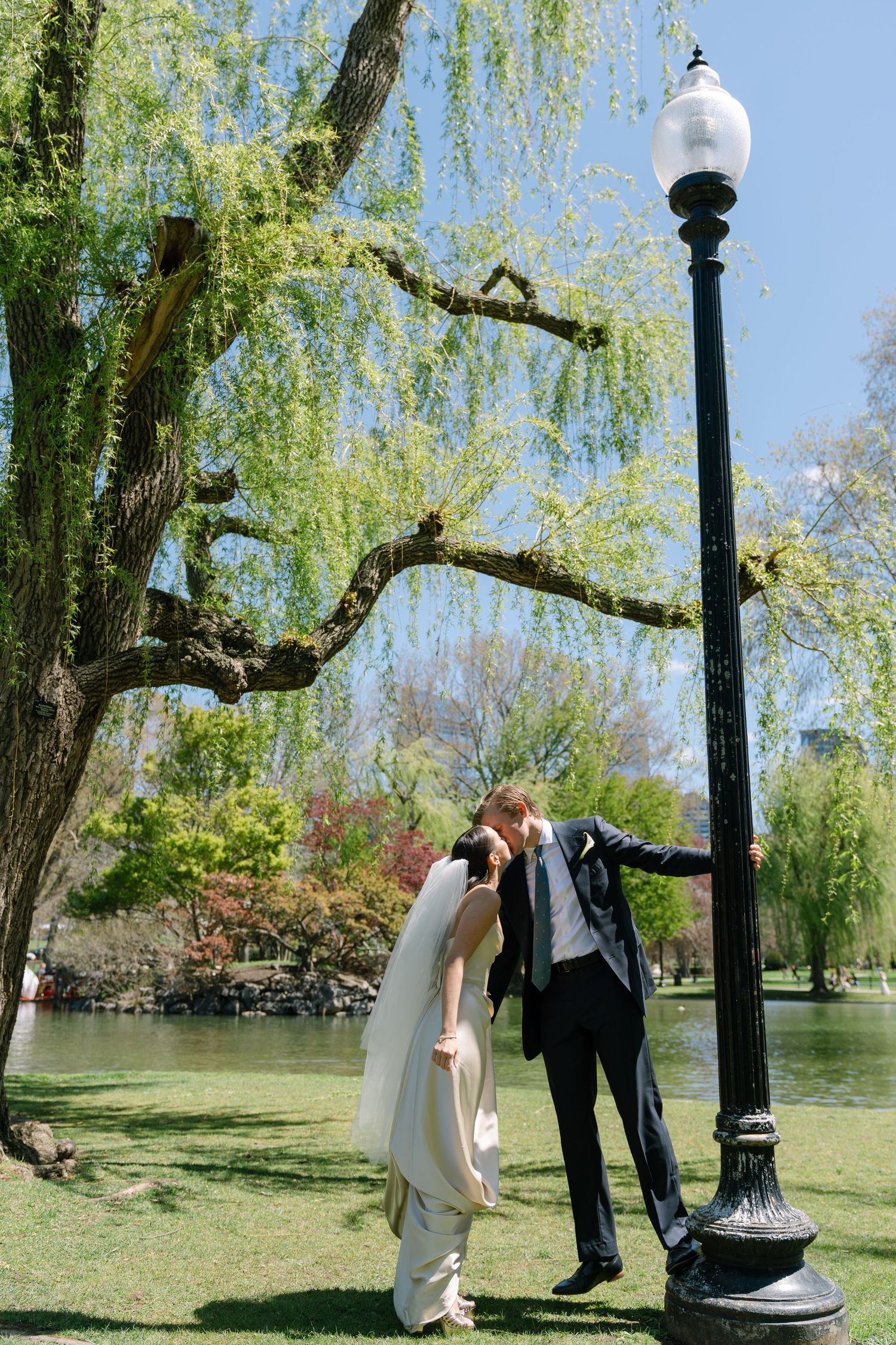 Bride and groom sharing a romantic kiss by a lamppost in Boston Public Garden with a weeping willow tree backdrop.
